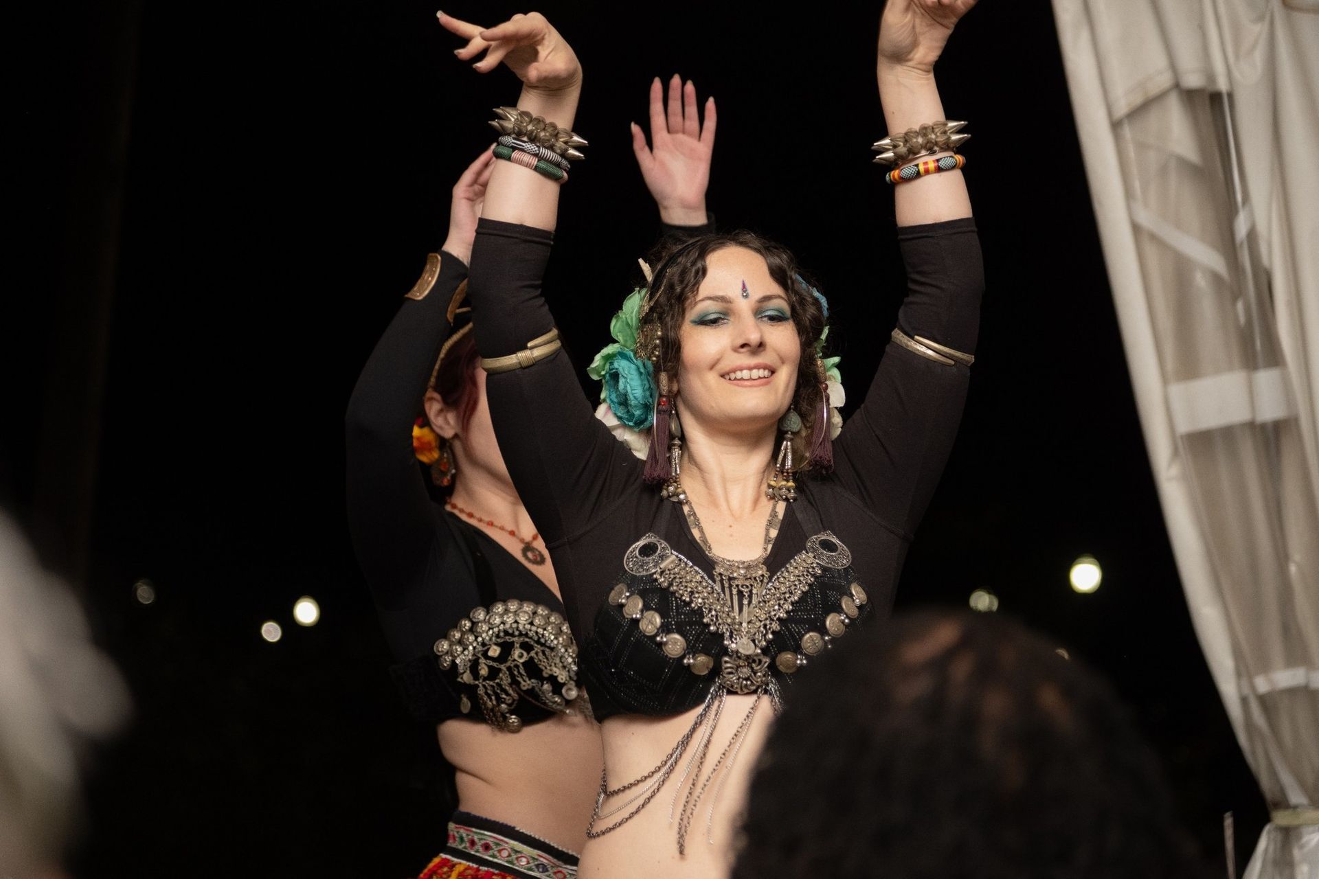 Two women performing belly dance outdoors at night, arms raised, smiling.