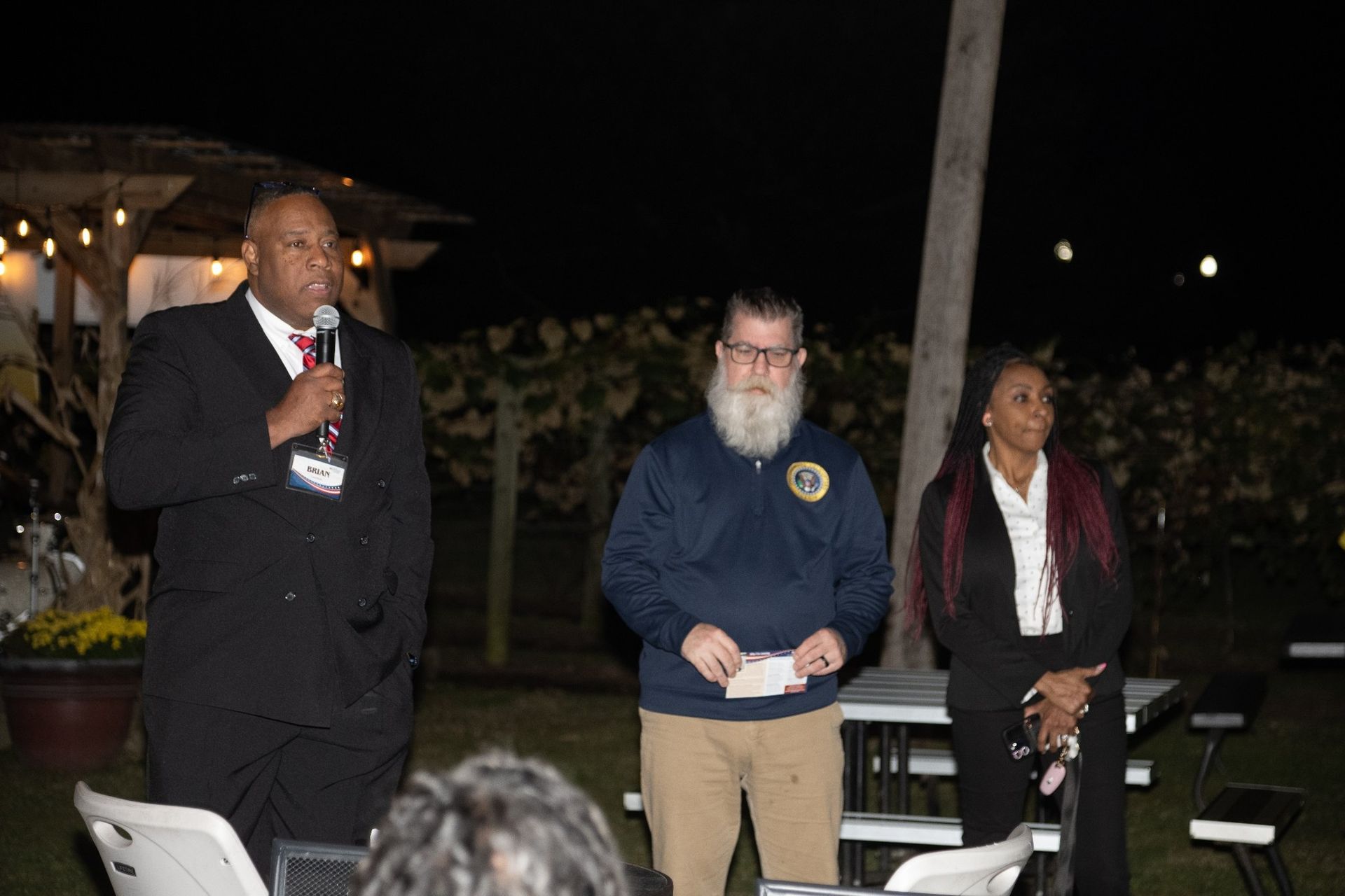 Three people standing outdoors. A man in a suit speaks into a microphone. Two others stand nearby. 