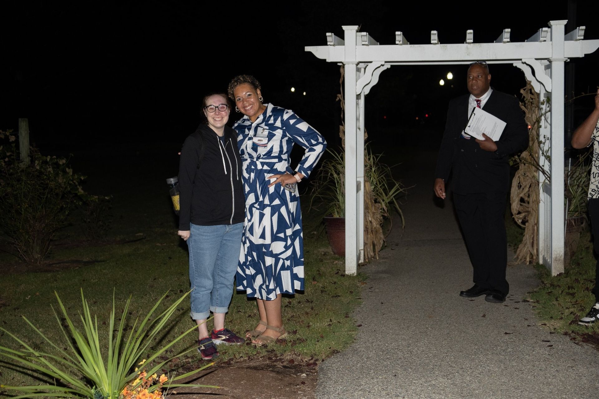 Woman in dress and woman in hoodie pose next to white arbor at night. Man in suit stands nearby.