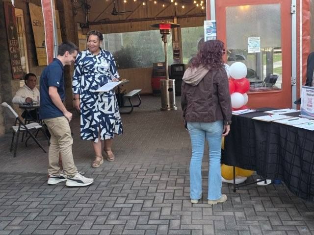 A person in a blue shirt bends over, speaking to a person in a patterned dress, at an outdoor event.