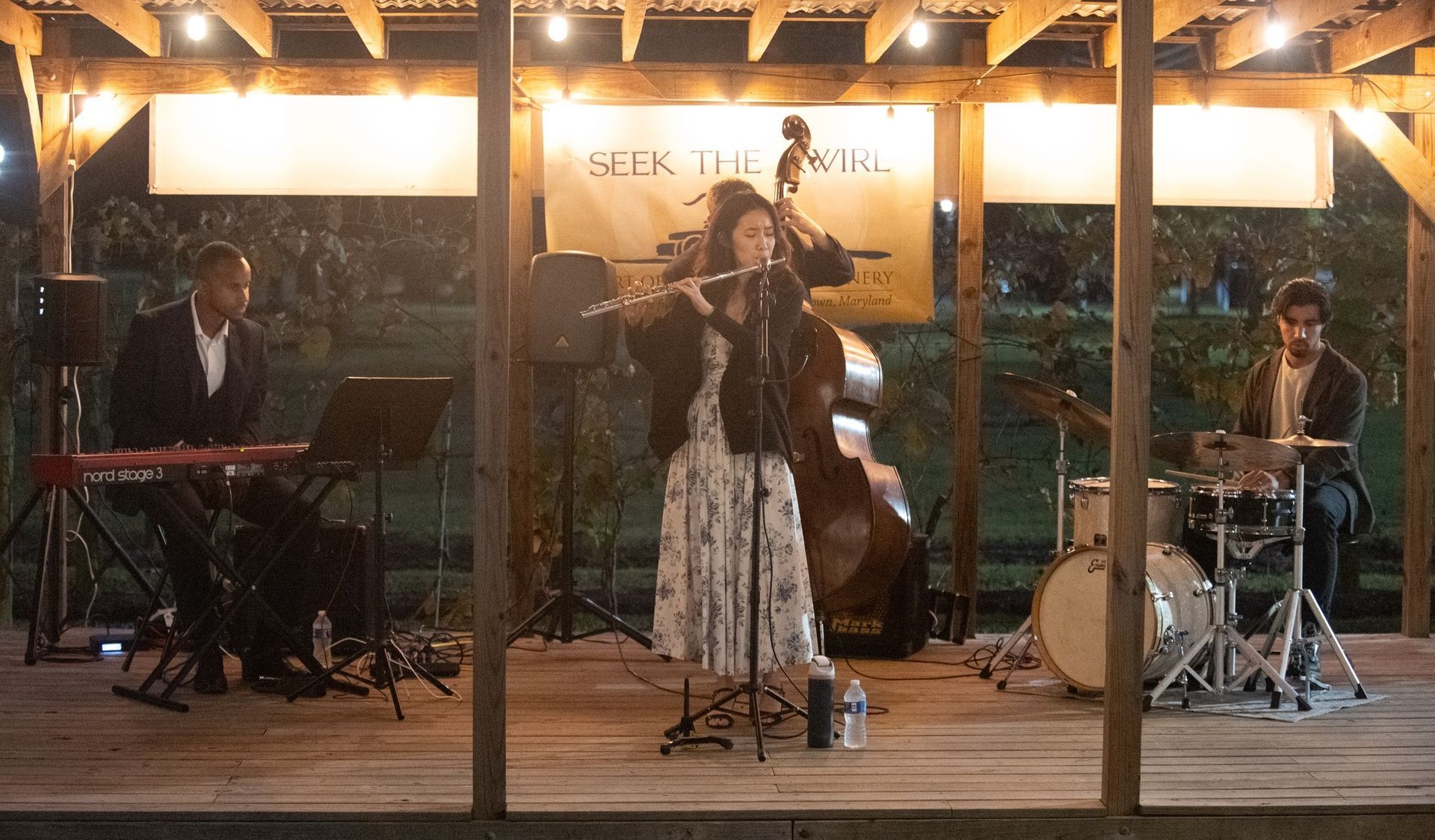 Jazz band performing outdoors at night: flute player center stage, drums, keyboard, and bass.