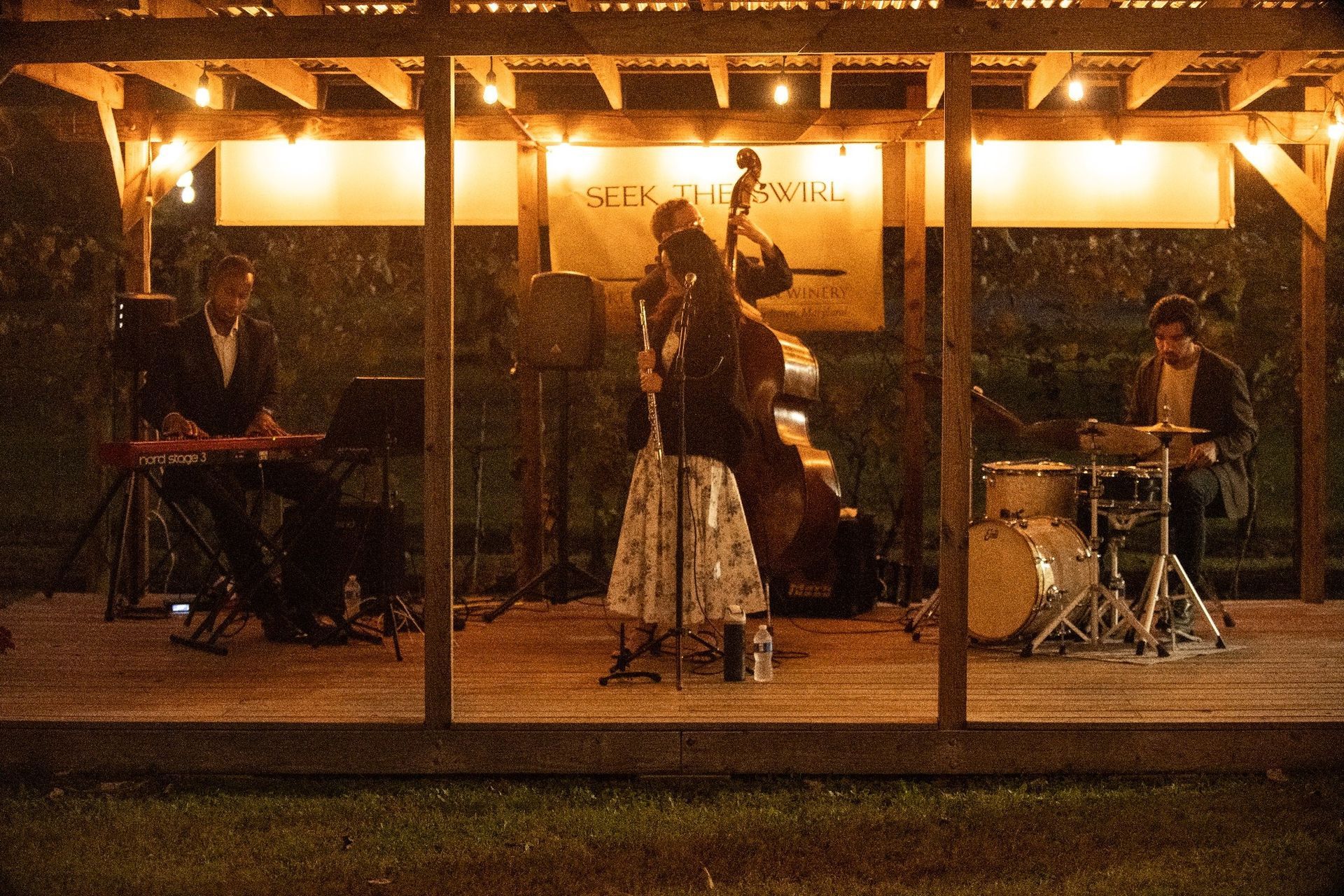 Jazz band performing on a lit outdoor stage at night; keyboard, bass, drums, and vocalist.