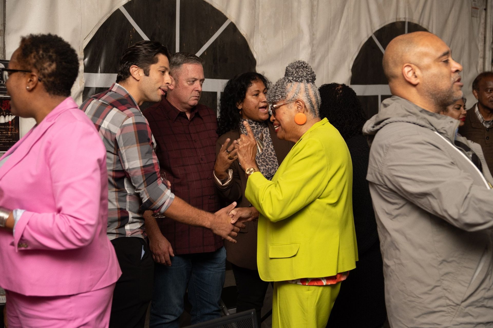 People greet each other at an event, one woman in lime green suit, others in casual attire, indoors.