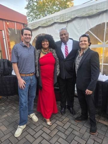 Four people pose outdoors. Woman in red dress stands between men in suits and casual attire.