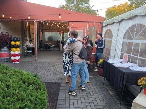 People hugging outdoors at a brick-paved event with a tent, tables, and string lights.