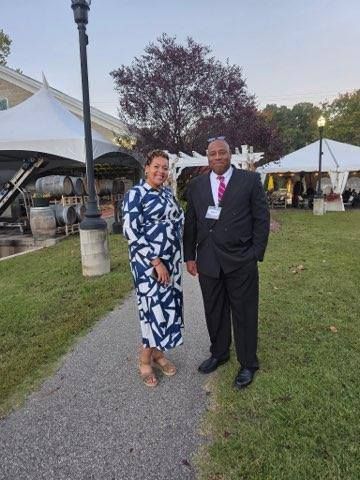 Woman in blue and white dress, man in a suit, standing outdoors near tents and a building.