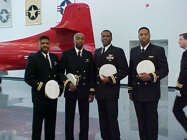 Four African American naval officers in dress uniforms stand in front of an aircraft. They each hold a white cap and smile.