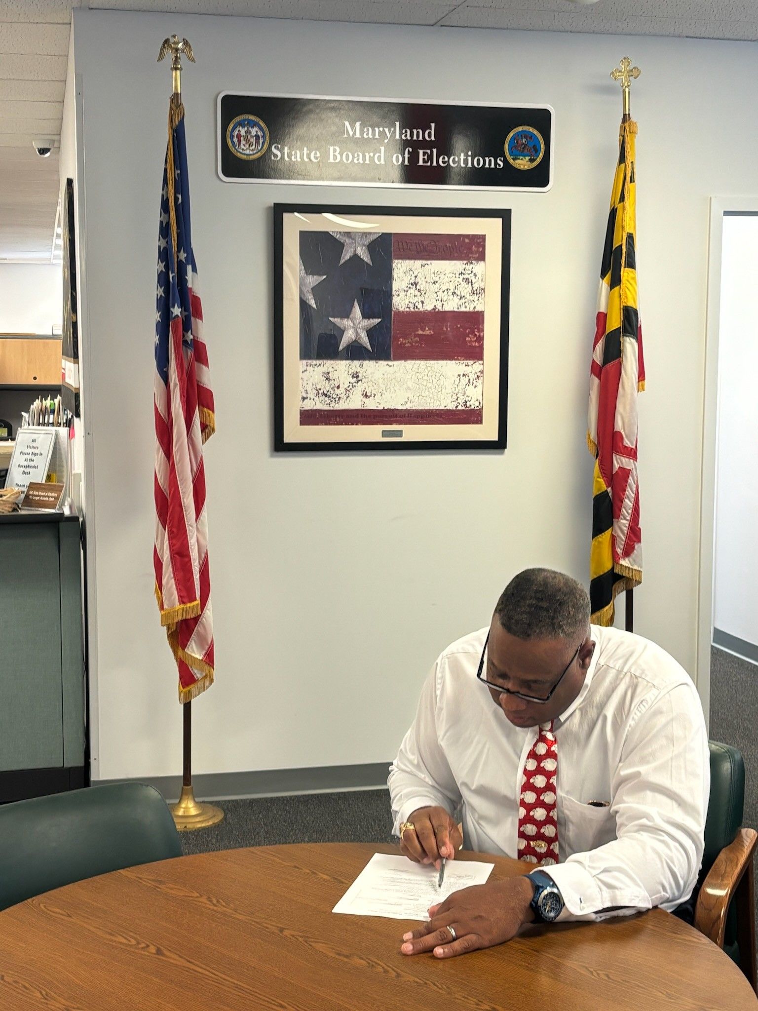 A man in a white shirt and tie sits at a table, signing documents. American and Maryland flags stand behind him.