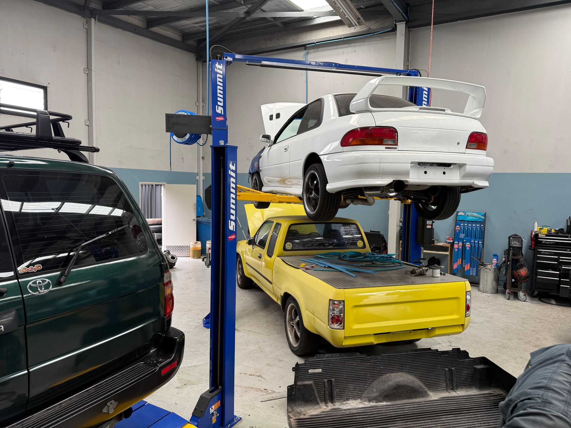 A White Subaru Impreza is Hoisted on a Vehicle Lift Above a Yellow Pickup Truck — Mad Monkeys Mechanical in Cairns City, QLD