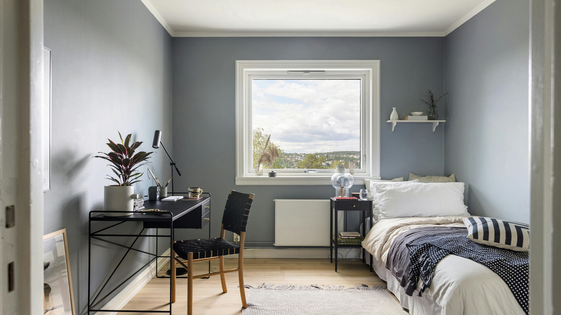 Small blue bedroom with desk, bed, and window overlooking a landscape.