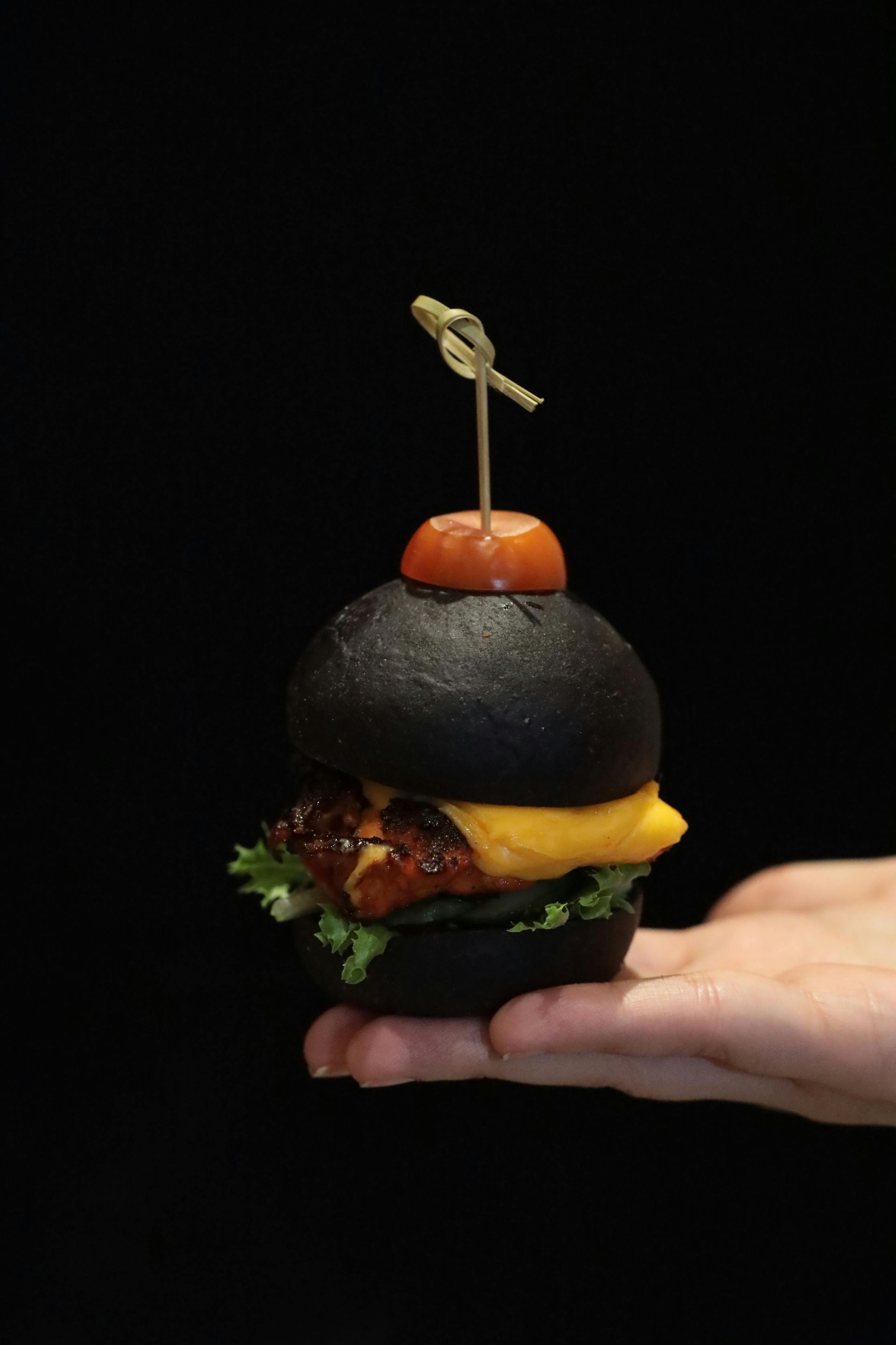 A hand holds a small black slider burger topped with a cherry tomato and a cocktail pick against a black background.