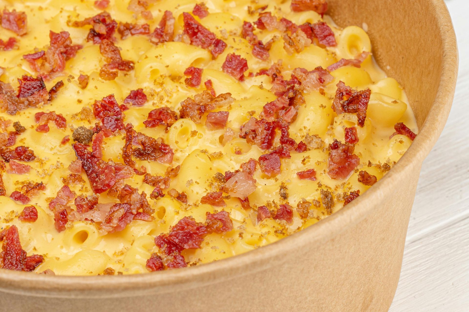 A close-up of macaroni and cheese topped with crispy bacon bits in a round, light-brown paper container.