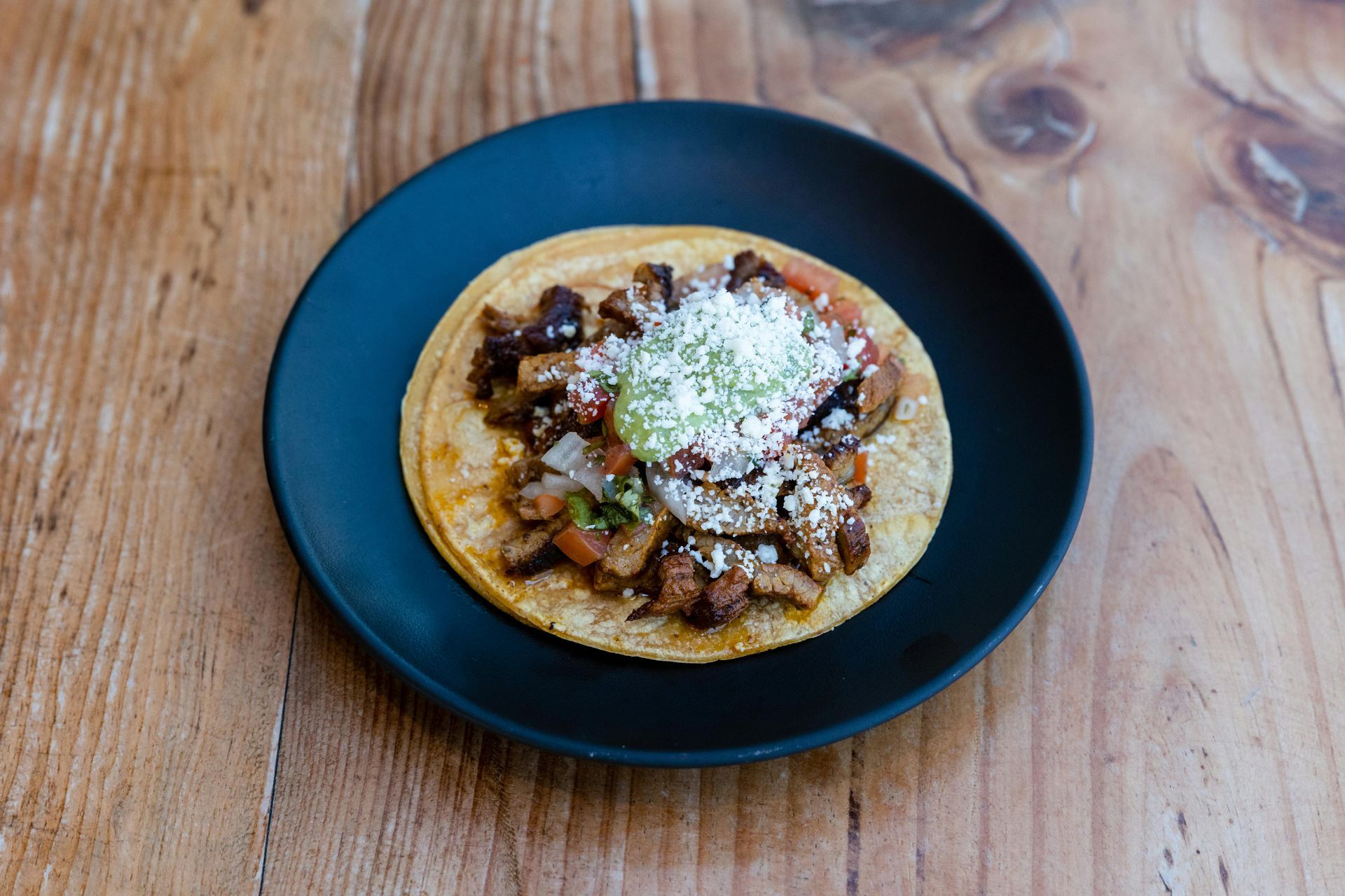 A single taco topped with meat, green salsa, and crumbled cheese on a black plate set on a wooden table.