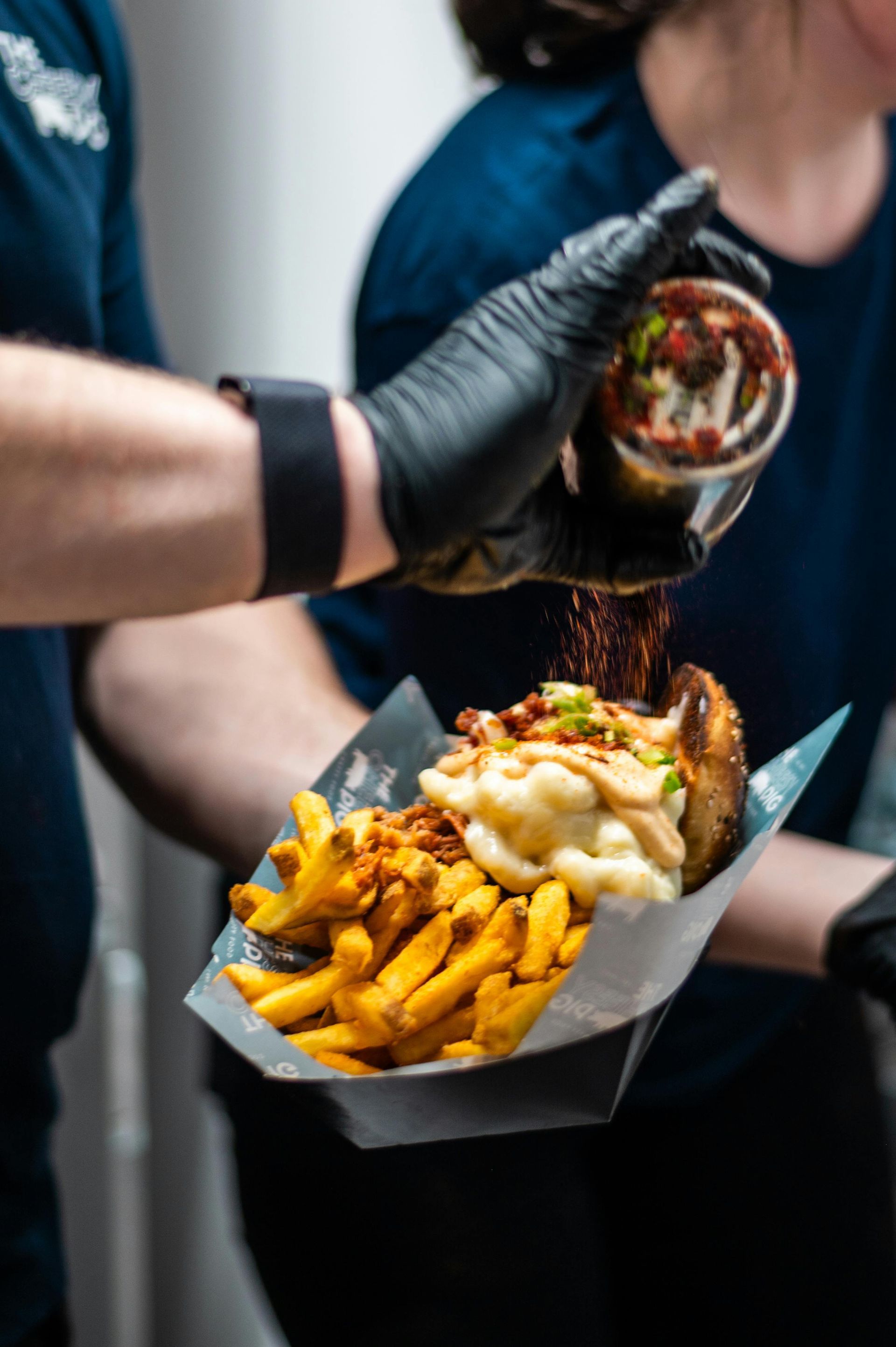 A person in black gloves sprinkles seasoning over poutine.
