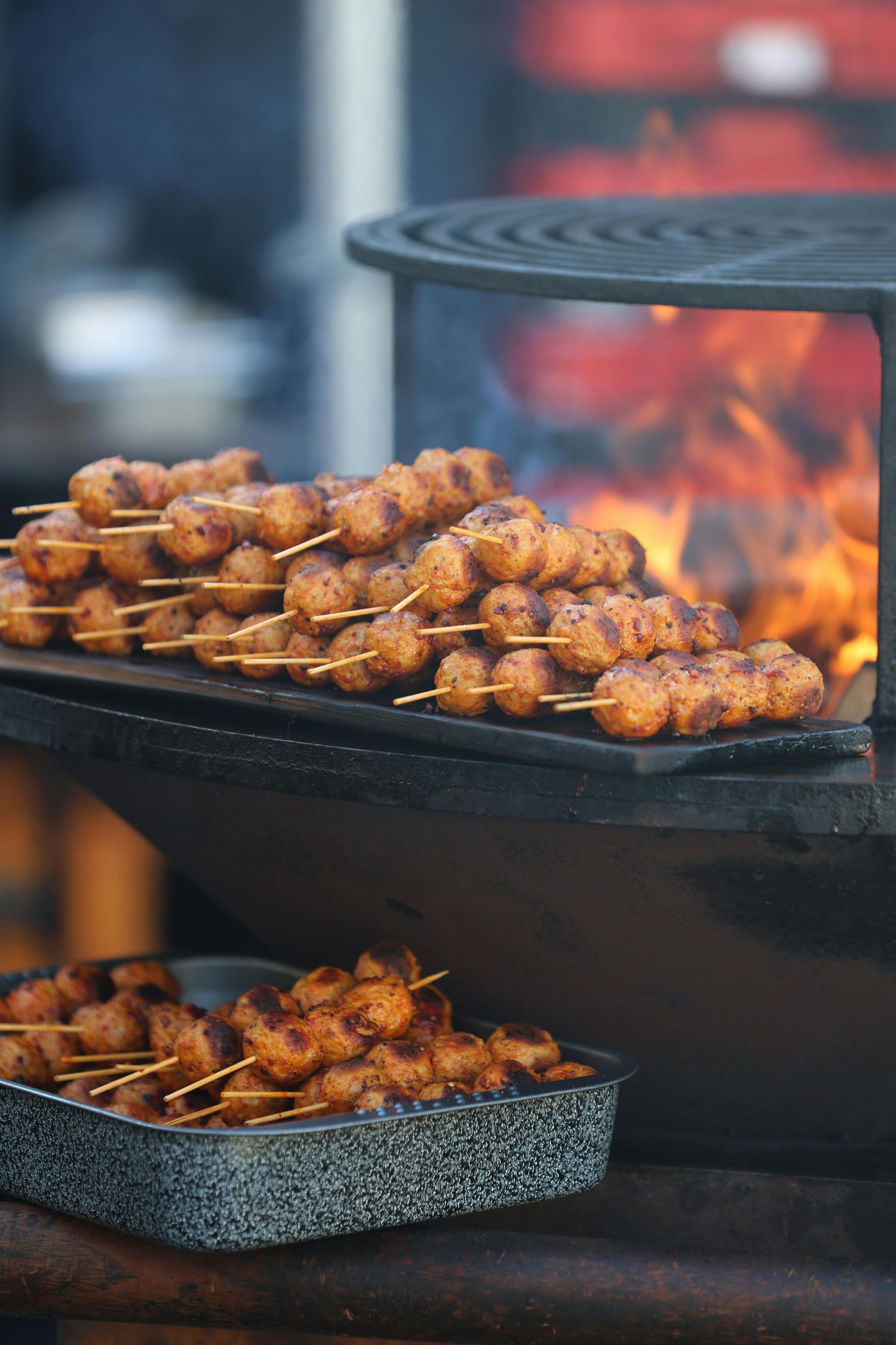 Skewered meatballs grilling on a barbecue fire with a tray of finished skewers below.