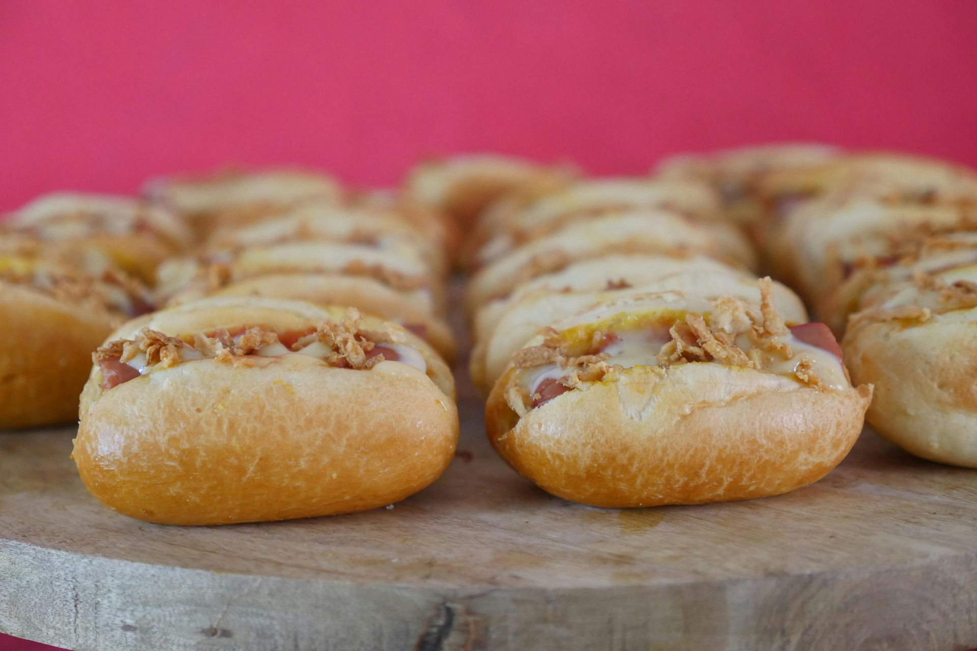 A tray of small, golden-brown philly cheese steak  sliders topped with crispy fried onions against a solid pink background.