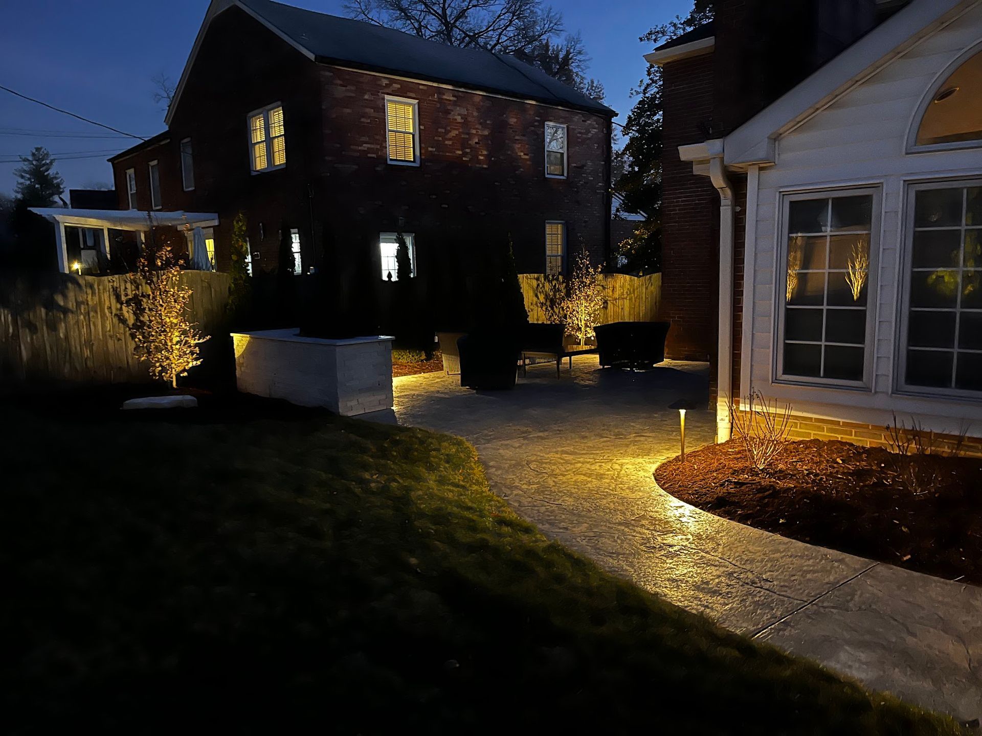 Exterior night view of a brick building and a white house with illuminated walkway. The scene is lit with spotlights.
