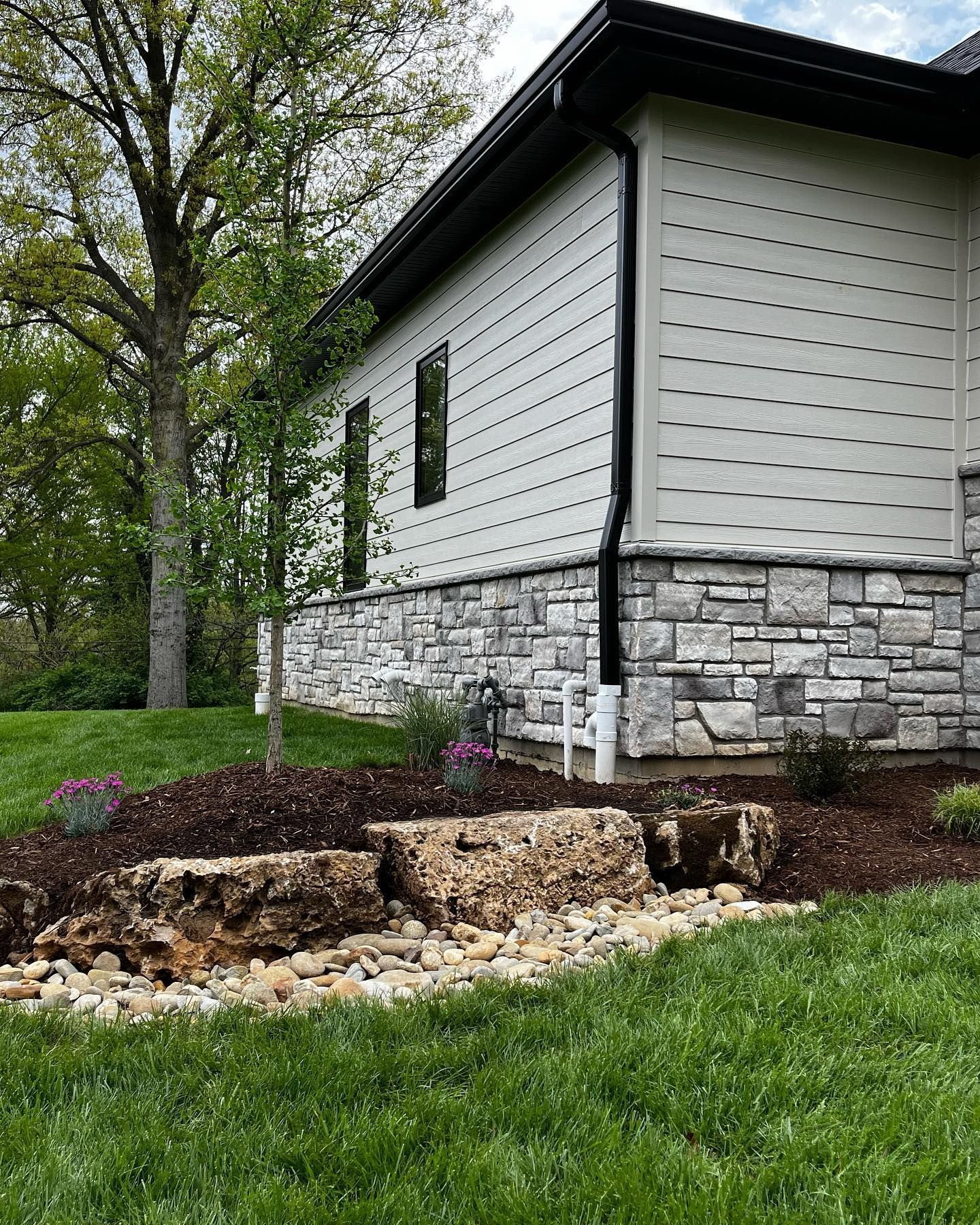 Exterior view of a modern house with stone veneer base, light siding, and a landscaped yard with rocks and plants.