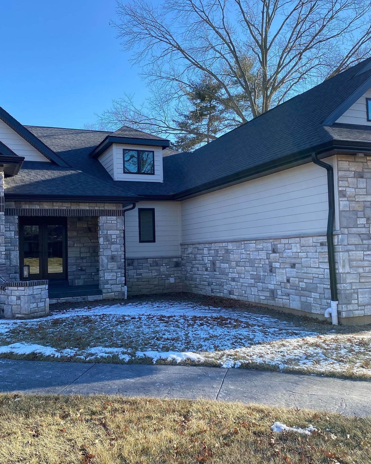 A house with gray stone and light siding. Snow covers the ground. Clear, blue sky.
