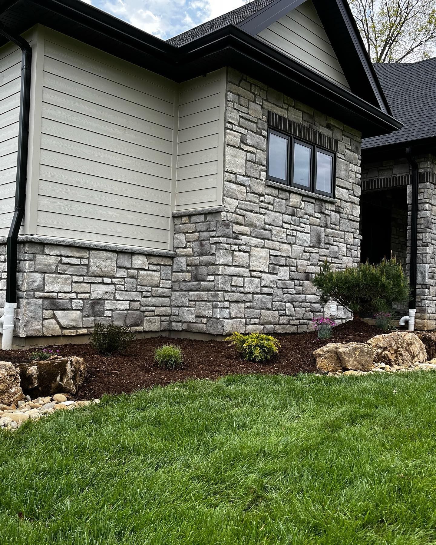 Exterior of a house with stone and light gray siding. Dark gutters and trim. Landscaping includes mulch, 