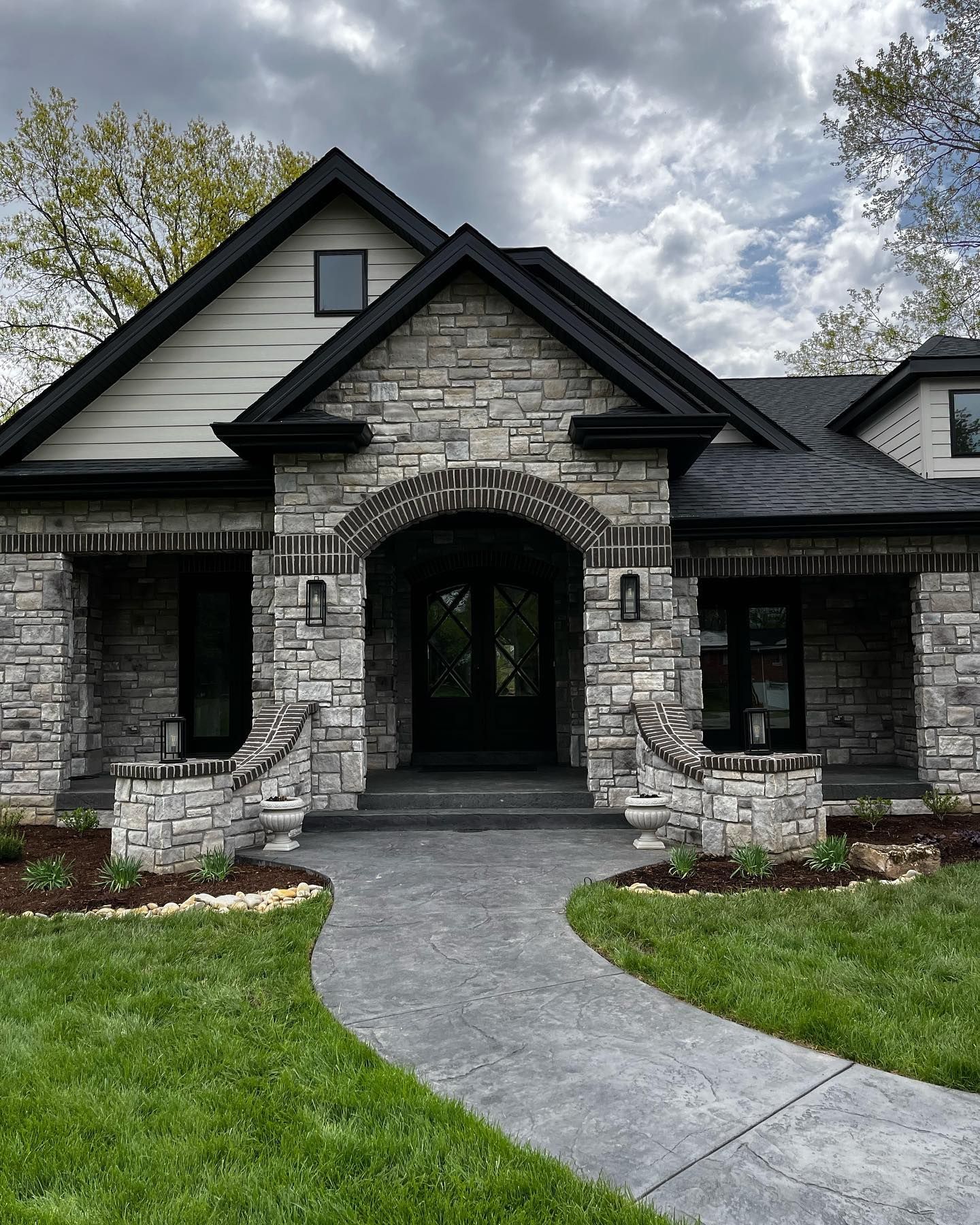 Stone facade house with a dark roof, entryway, and walkway. Green lawn and overcast sky.