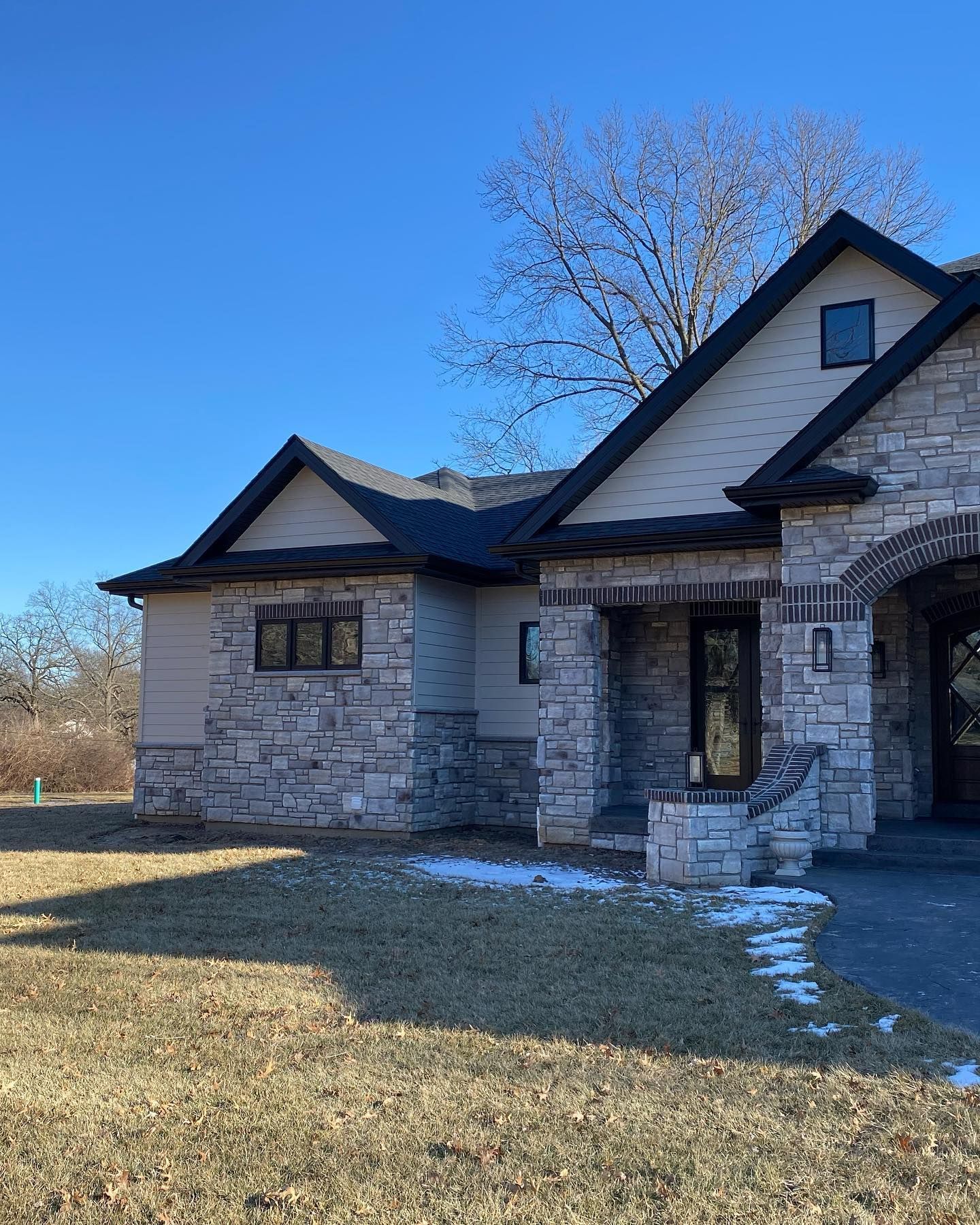 A modern house with stone and tan siding against a clear blue sky. The architecture features dark trim and small windows.