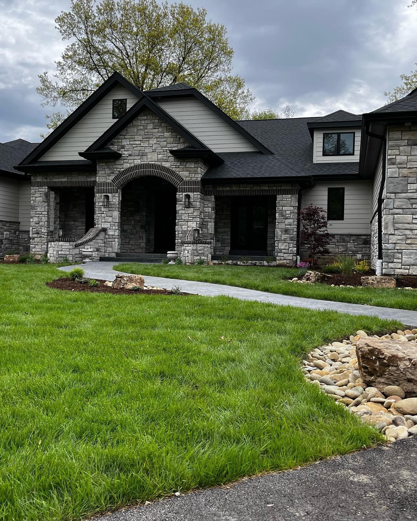 A modern, stone-faced house with a dark roof and green lawn under an overcast sky. A walkway leads to the front entrance.