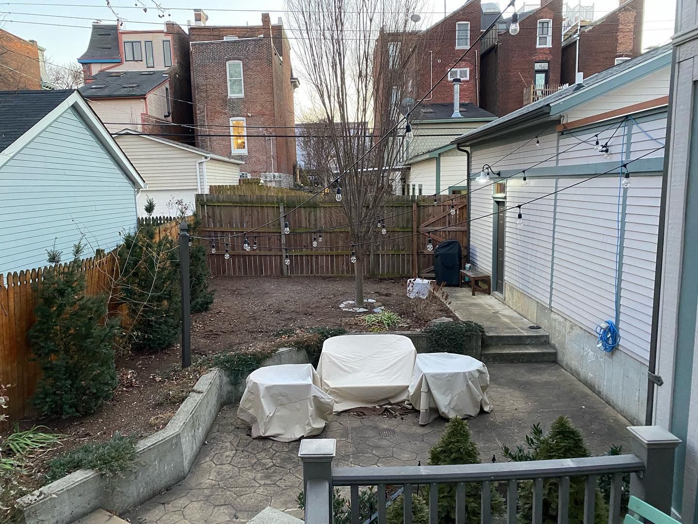 Backyard with a concrete patio, covered furniture, a wooden fence, and surrounding brick buildings.  Overcast sky.
