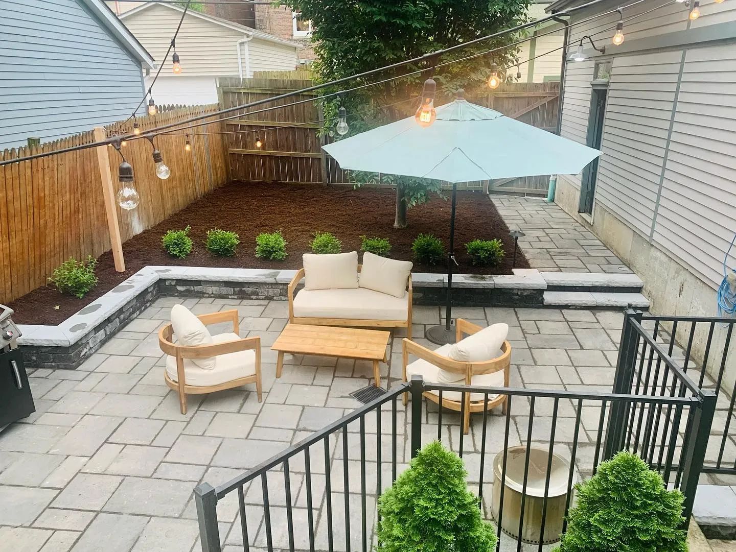 Cozy backyard patio with light blue umbrella, wooden furniture, string lights, and a fenced-in garden area.