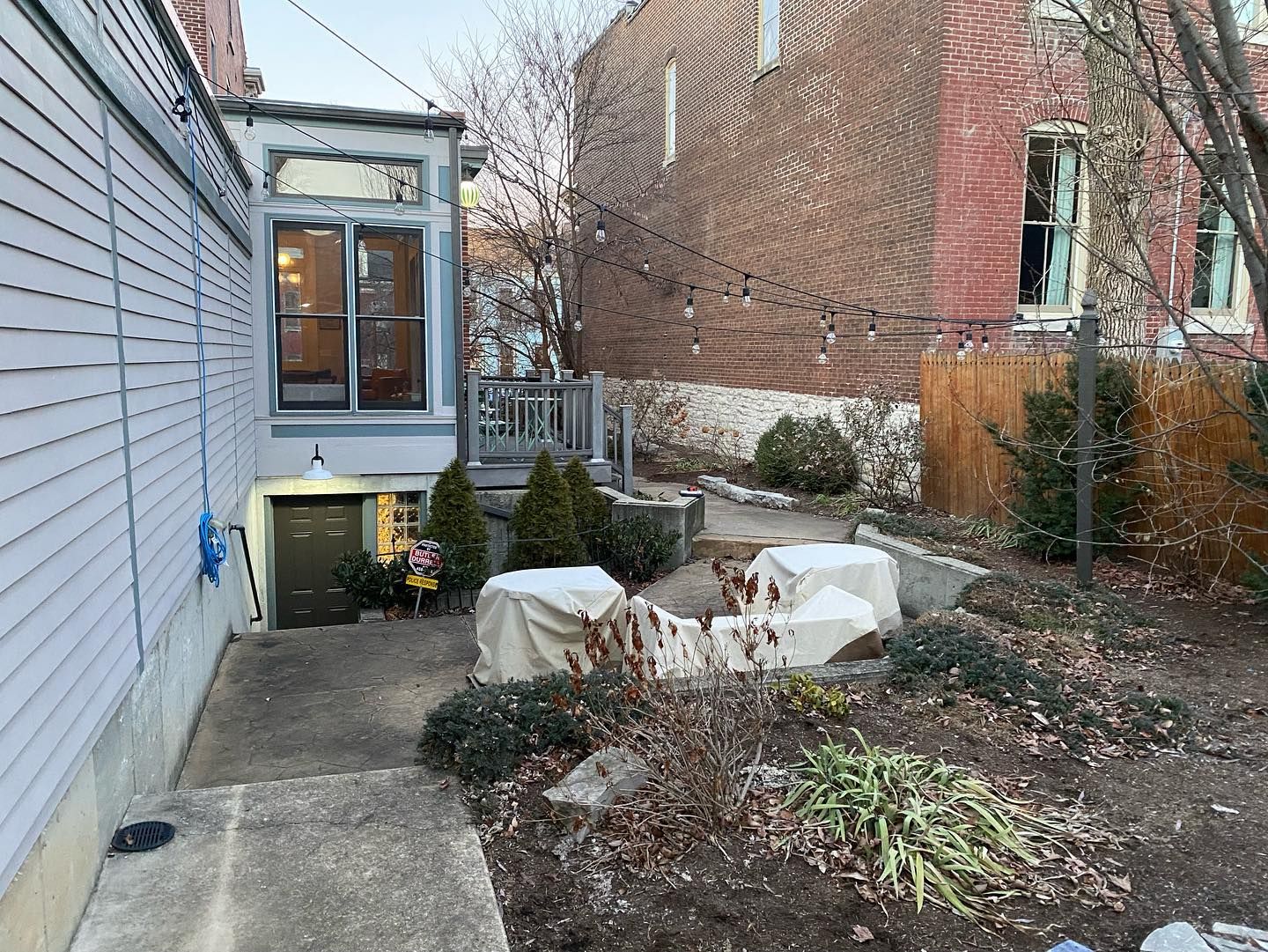 Backyard with a concrete path, garden beds, covered seating, and a light-strung fence. 