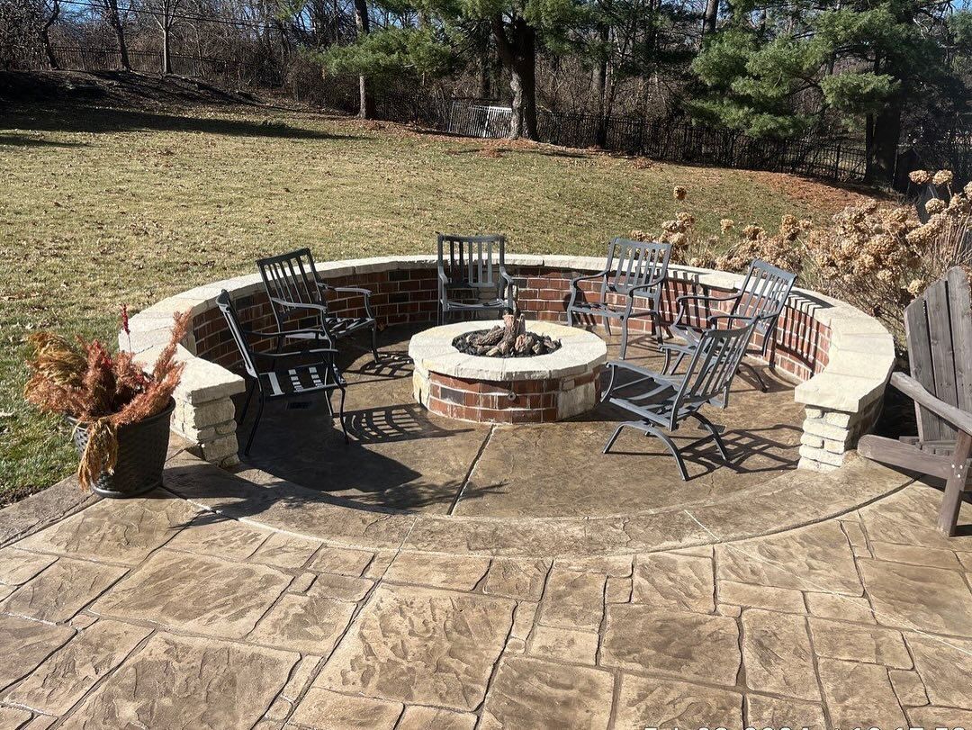 Circular fire pit area with brick fire pit, stone wall, and chairs on a stamped concrete patio.