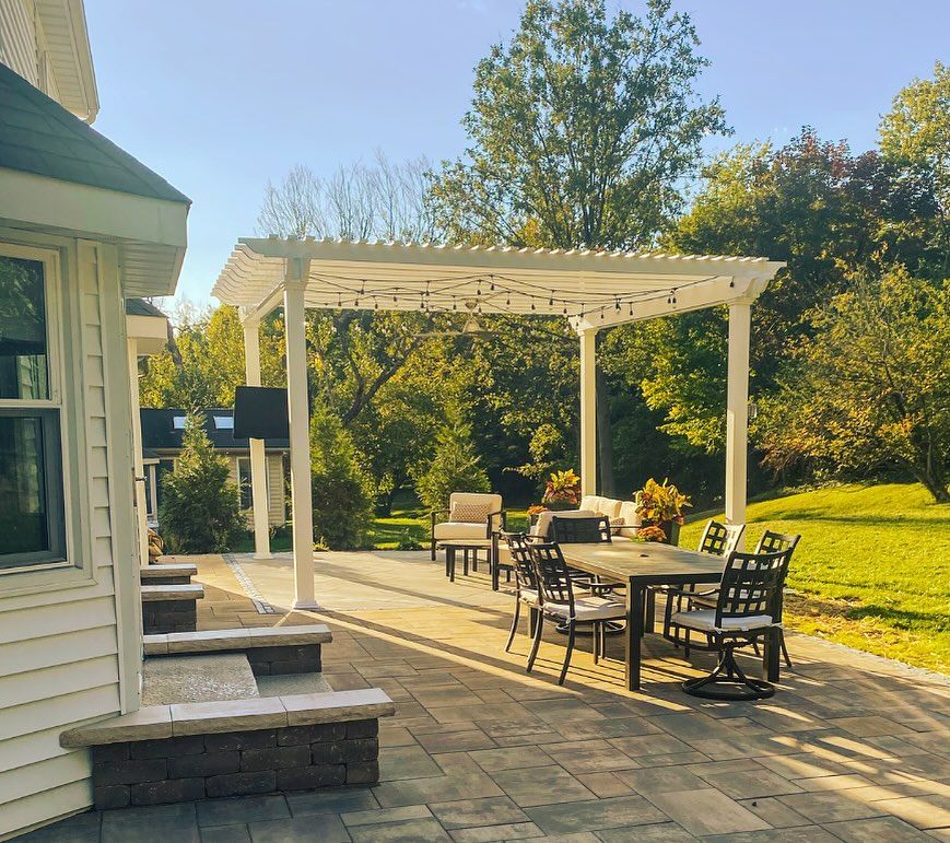 Outdoor patio with a pergola, table, chairs, and a grassy backyard. White structure with string lights, stone patio, 