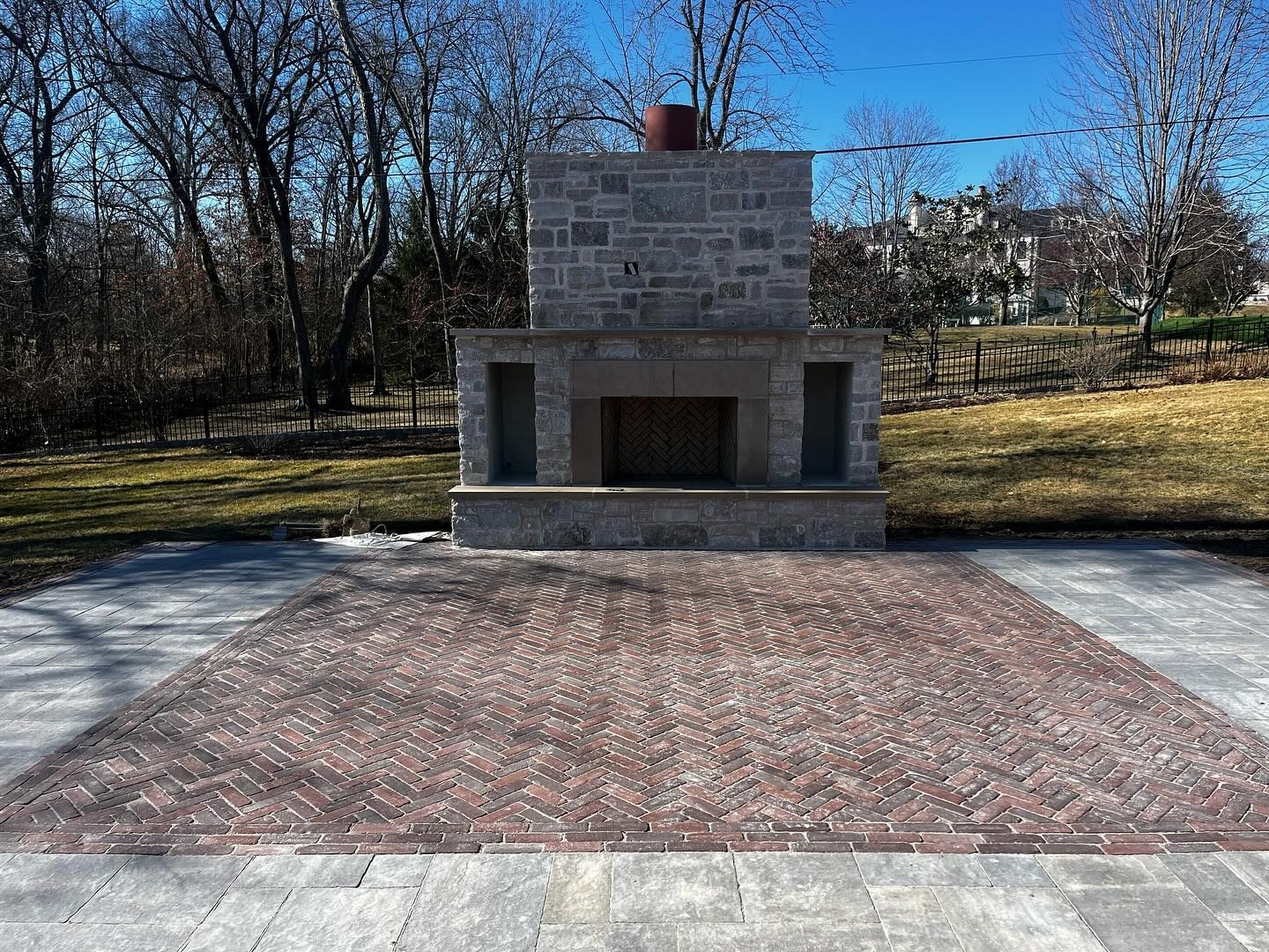 Outdoor stone fireplace on a brick patio. Trees and a grassy yard are in the background.