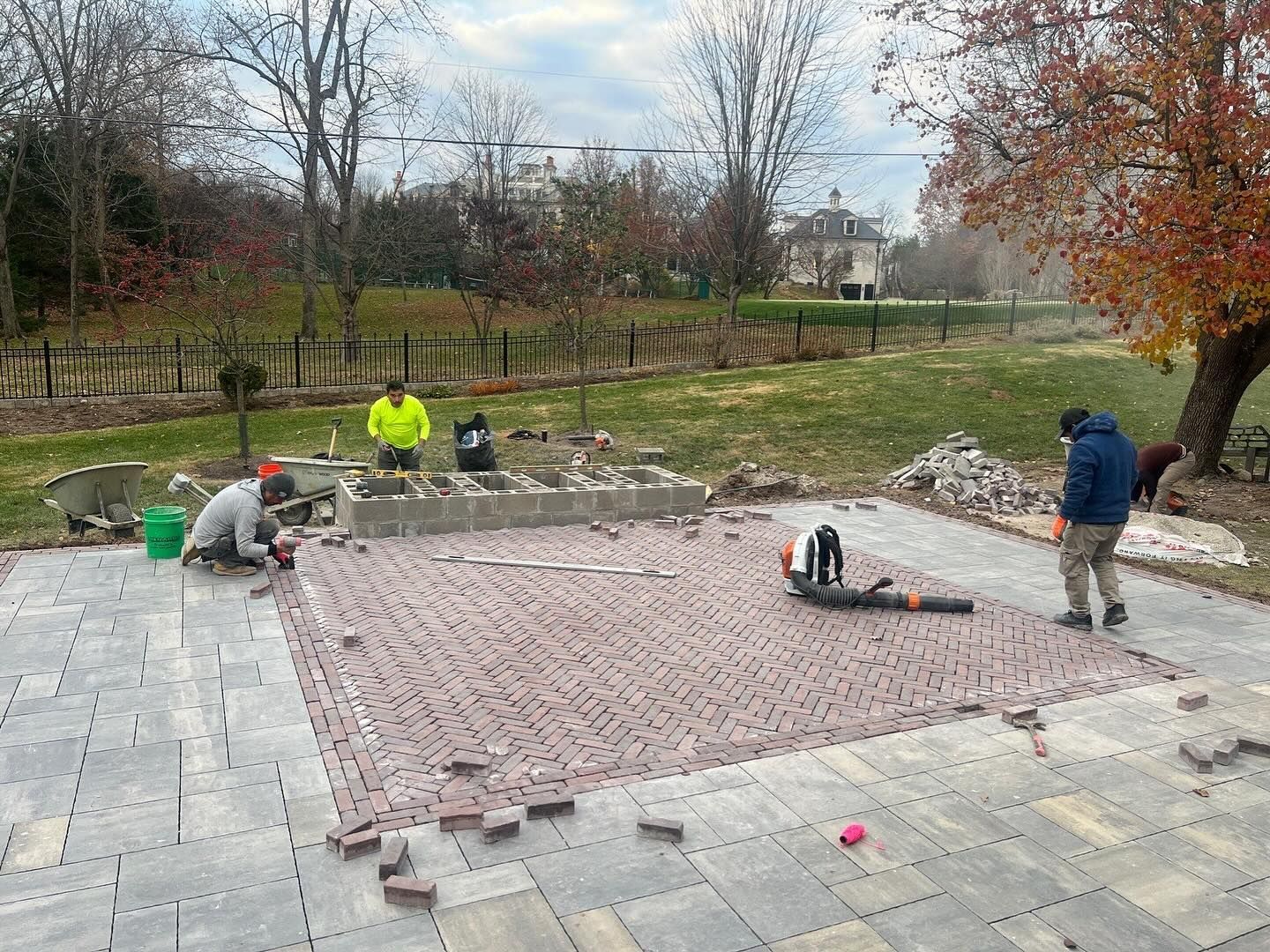 Construction workers laying brick pavers on a patio. Two workers use tools, while others oversee and sweep.