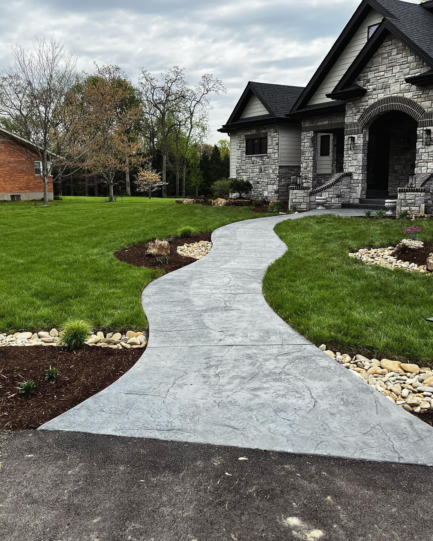 Curving gray concrete walkway leading to a stone-clad house with an arched doorway, set in a green lawn.