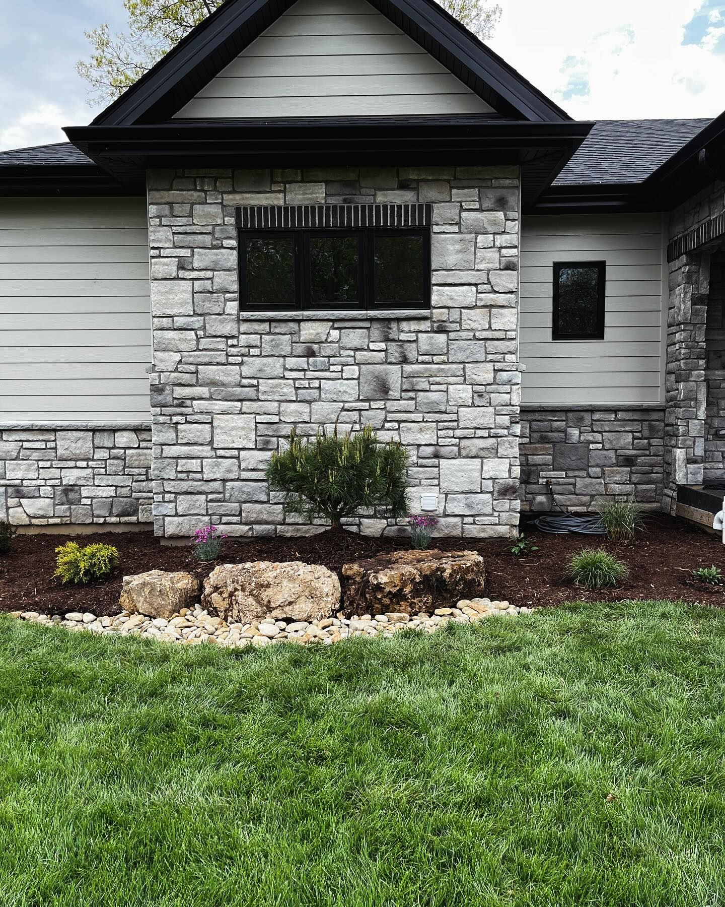 Stone-clad home exterior with gray siding and black trim. Landscaping includes rocks, mulch,