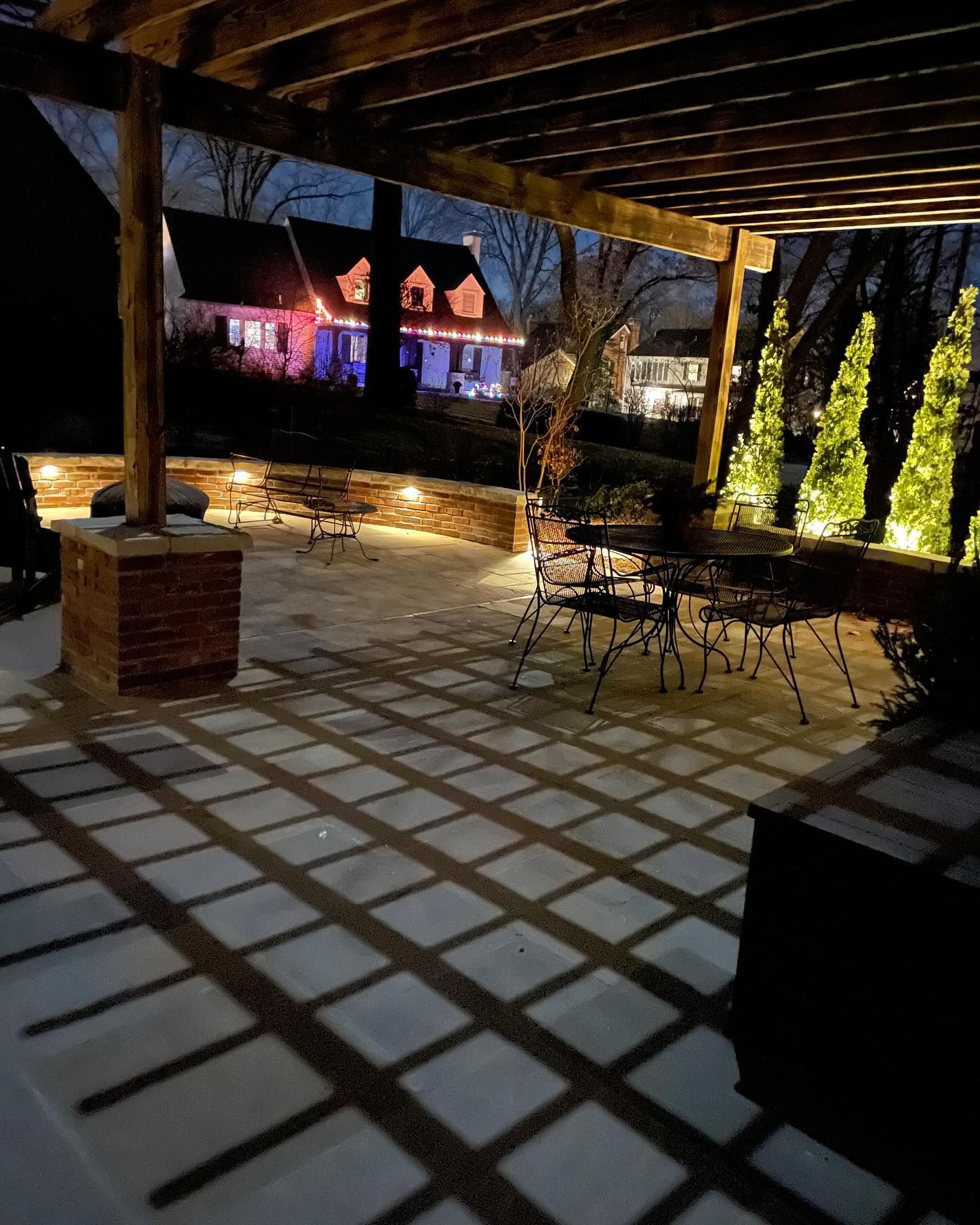 Nighttime patio with pergola and shadows, illuminated by built-in lights. In the background, a house with Christmas lights.