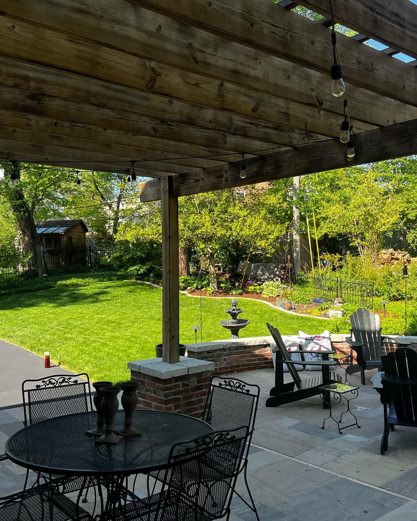 A backyard patio with a dining table and chairs under a wooden pergola, overlooking a green lawn and garden.