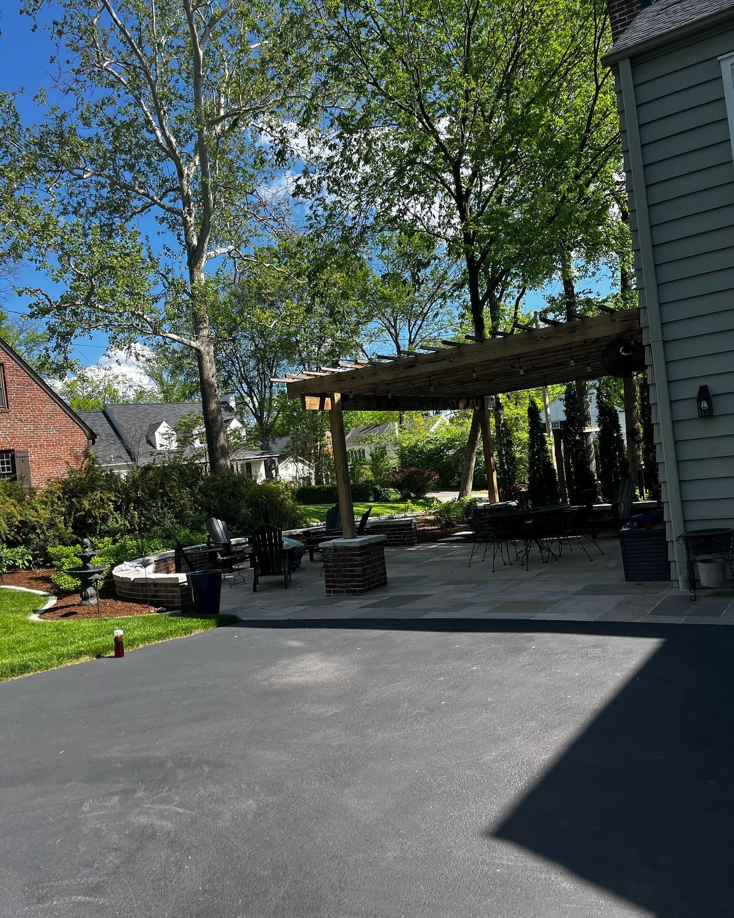 Asphalt driveway leading to a stone patio with a pergola, tables, and chairs. Trees and a house flank the scene.