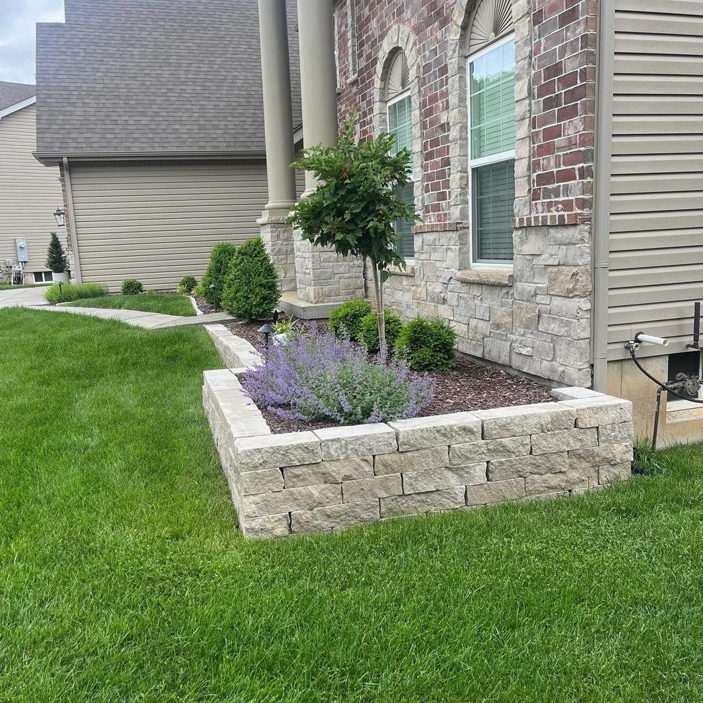 A manicured front yard with a raised stone flower bed filled with plants in front of a brick house.