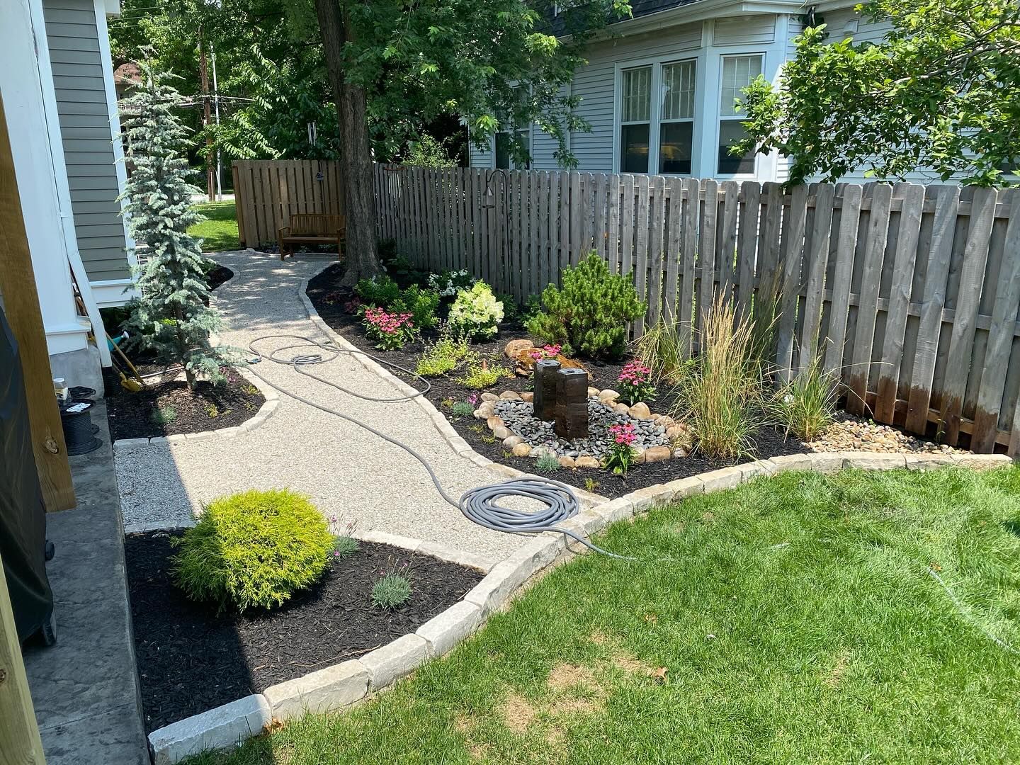 A landscaped path with a gravel surface winds through a garden bed bordered by a wooden fence and lawn.