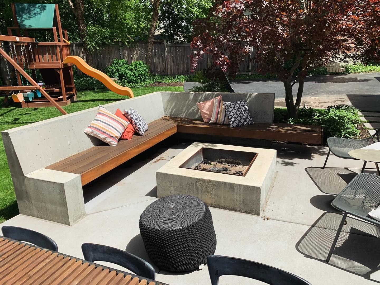Outdoor seating area with concrete benches, fire pit, and a wooden table.  A playground and tree are in the background.