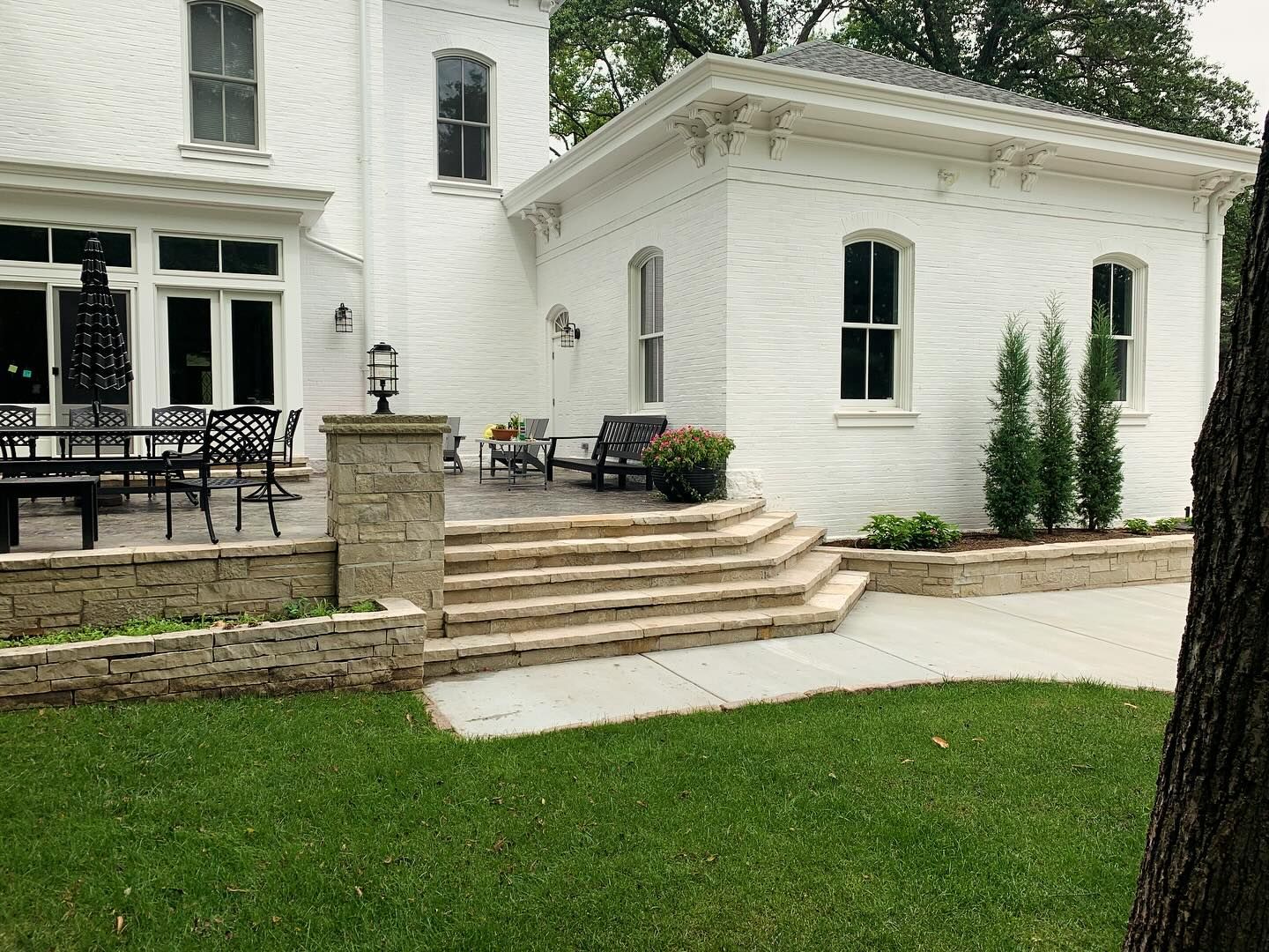 White brick home exterior with a patio and stone steps. Green grass, trees, and outdoor seating are present.