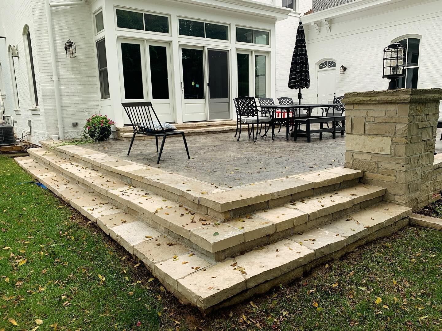 A stone patio with steps leading to a seating area with a table and chairs, beside a white house.