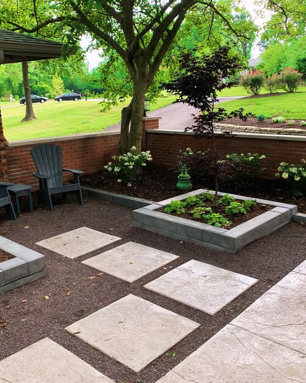 A backyard patio with square stepping stones, a raised garden bed, seating, and a brick wall, shaded by trees.