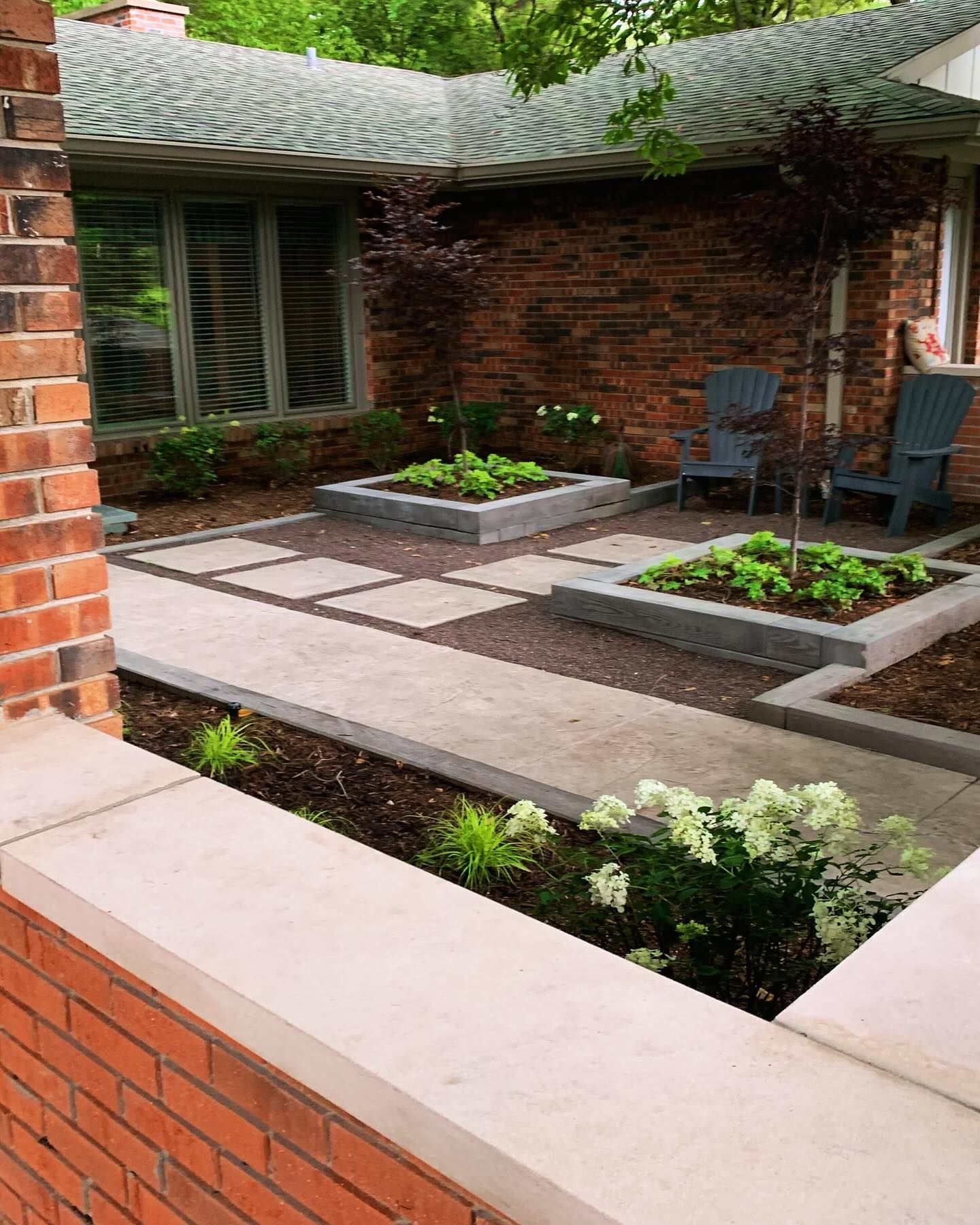 A brick house with a concrete walkway and raised garden beds. Two blue chairs sit on a patio, surrounded by greenery.