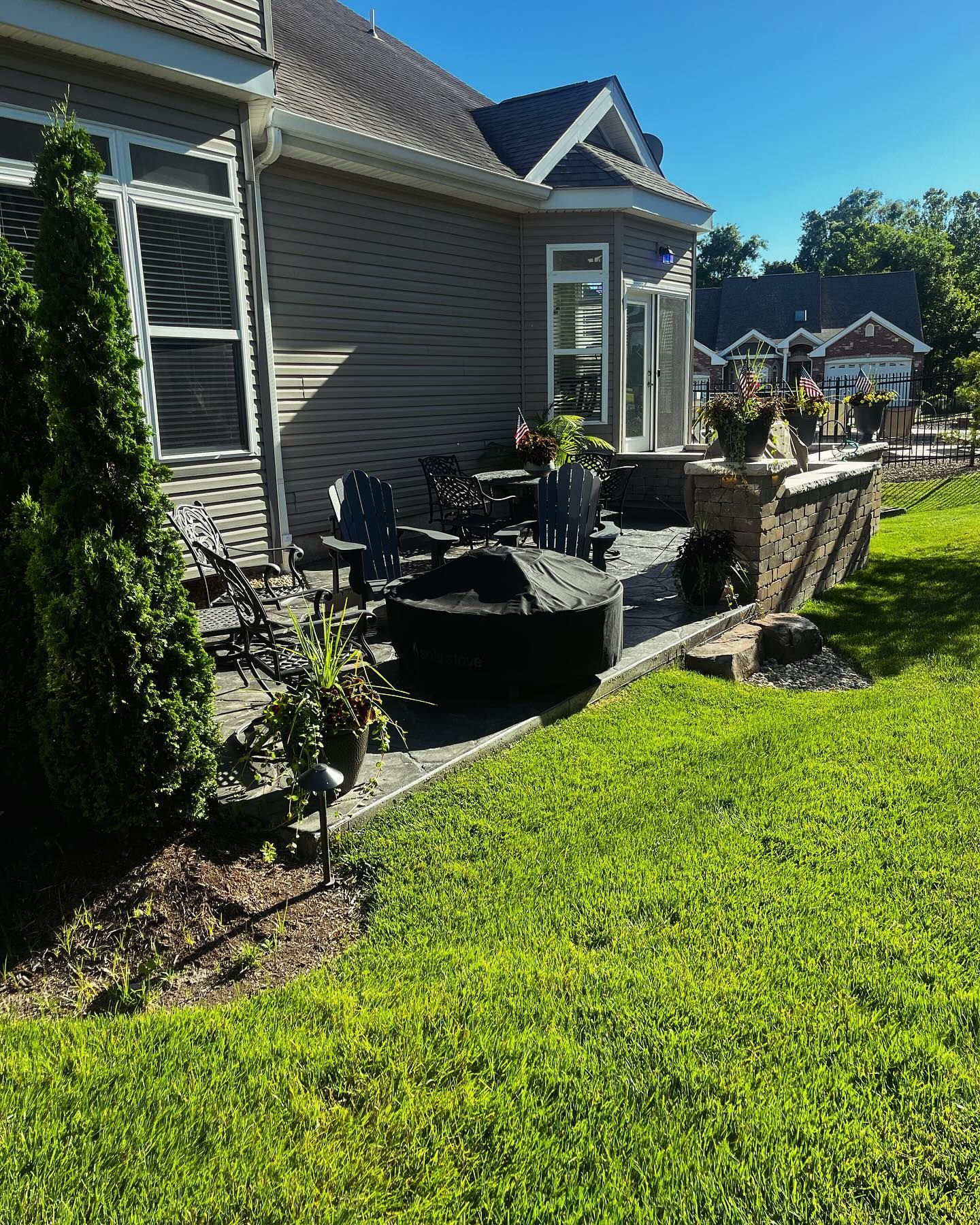A backyard with a deck, retaining wall, and house. Green grass surrounds the raised deck with a fire pit and patio furniture.