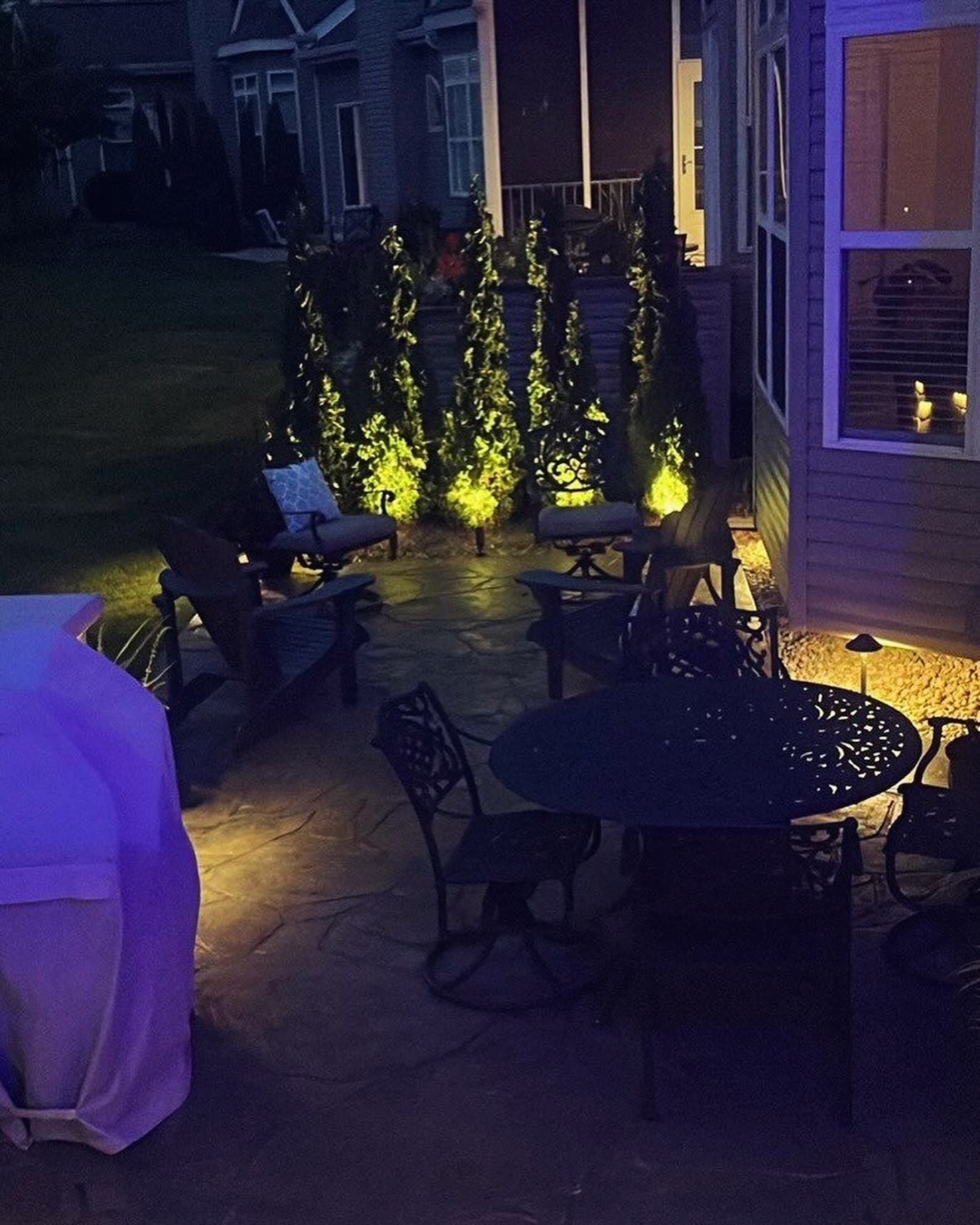 Nighttime patio with illuminated trees, seating, and a covered grill. Soft yellow lights highlight the space.