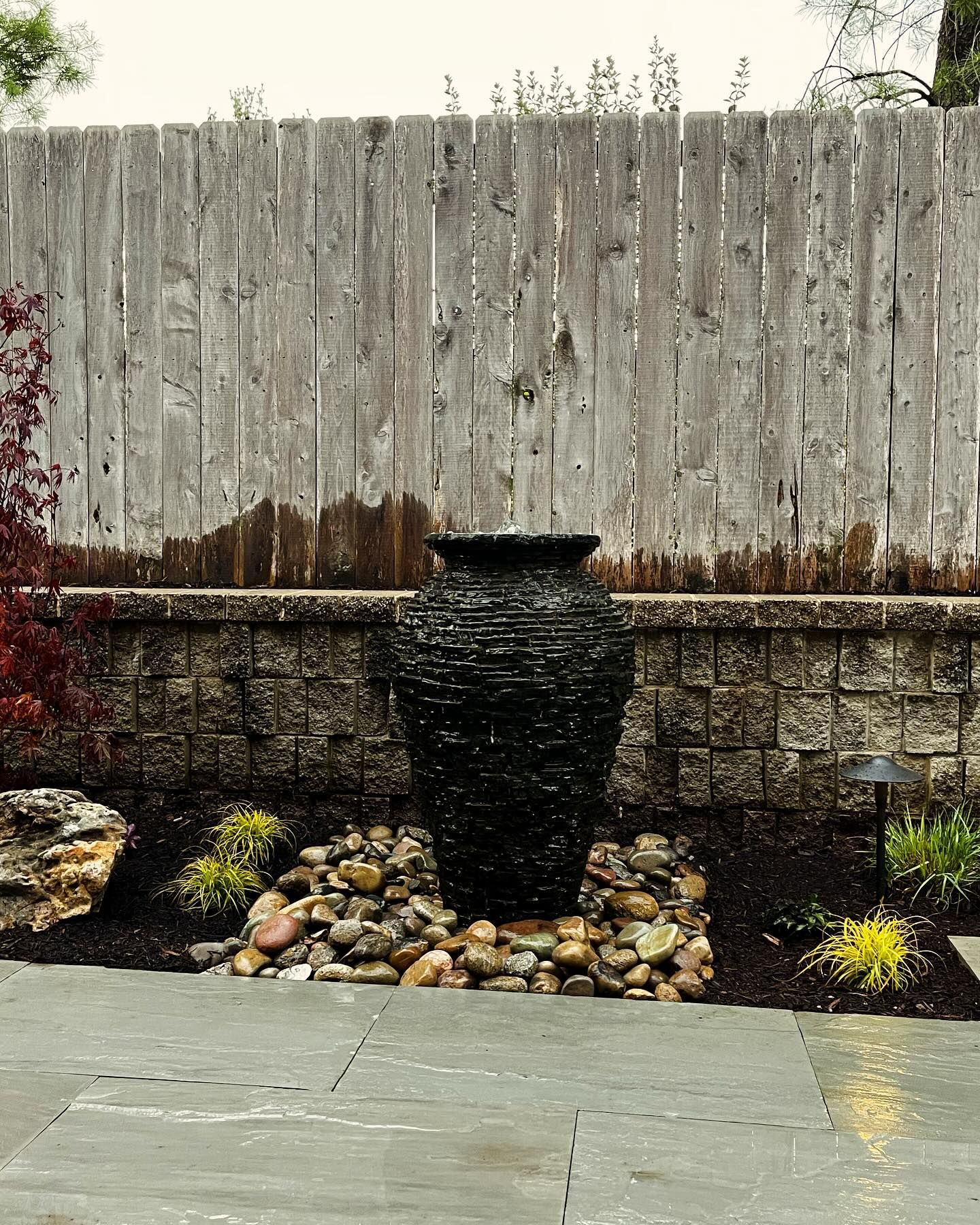 A black textured water fountain in the shape of a vase sits on a bed of rocks in front of a wooden fence.