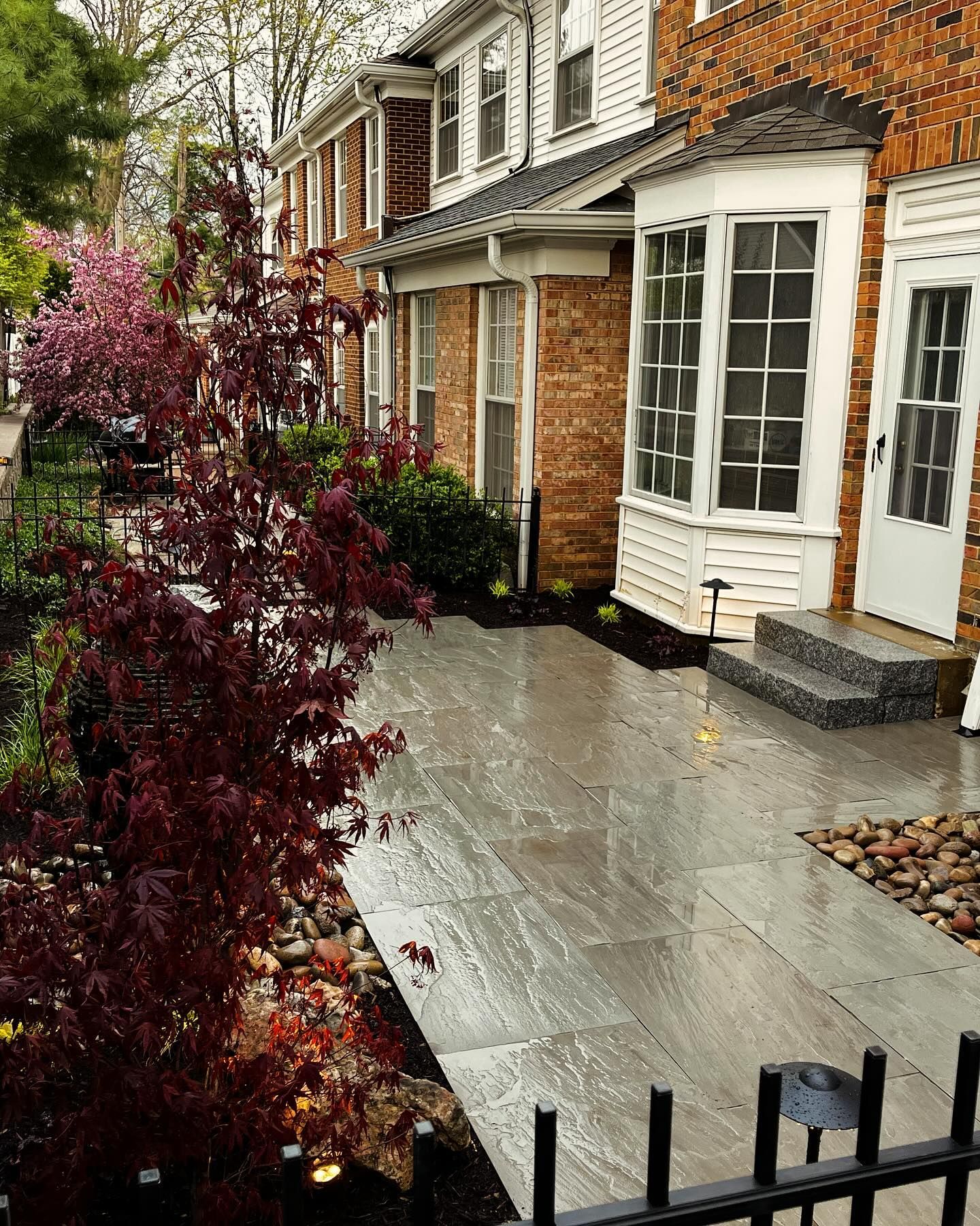 Brick townhouse with a gray stone patio lined with a dark red Japanese maple. A wrought iron fence borders the patio.